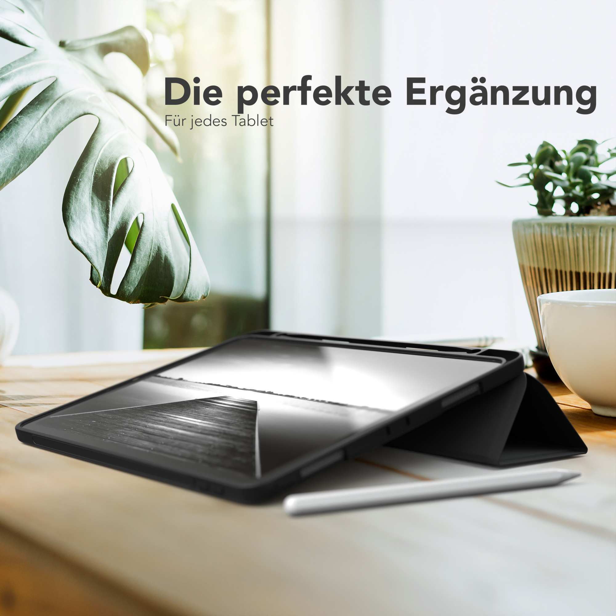 Tablet in schwarzem Case, Bleistift und Pflanze auf Holztisch. Fenster im Hintergrund.