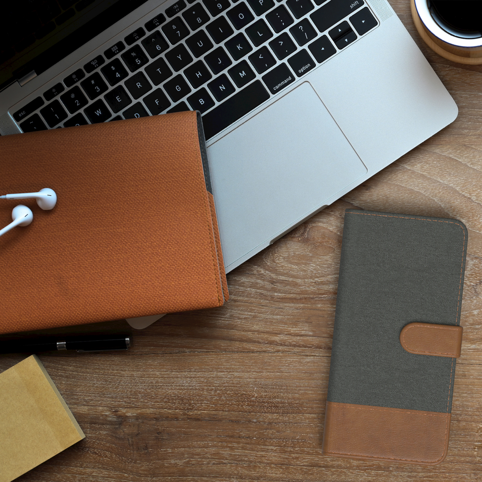 Ein Holzschreibtisch mit Laptop, Handyhüllen, Kopfhörern und Kaffeetasse. Stift und Notizblock sind ebenfalls vorhanden.