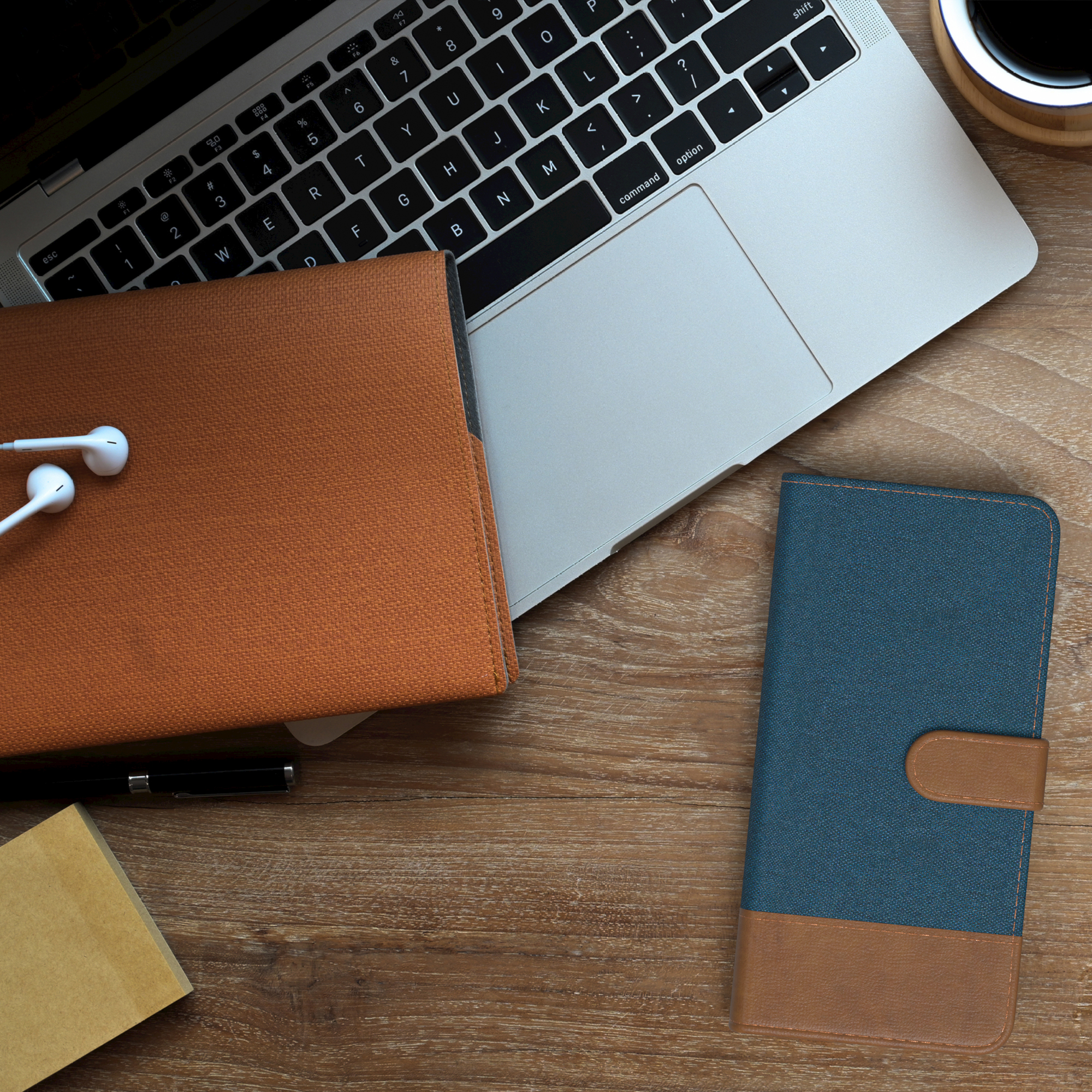 Laptop, Notizbuch, blau-braune Handyhülle, weiße Kopfhörer, Kaffeetasse, Holztisch.