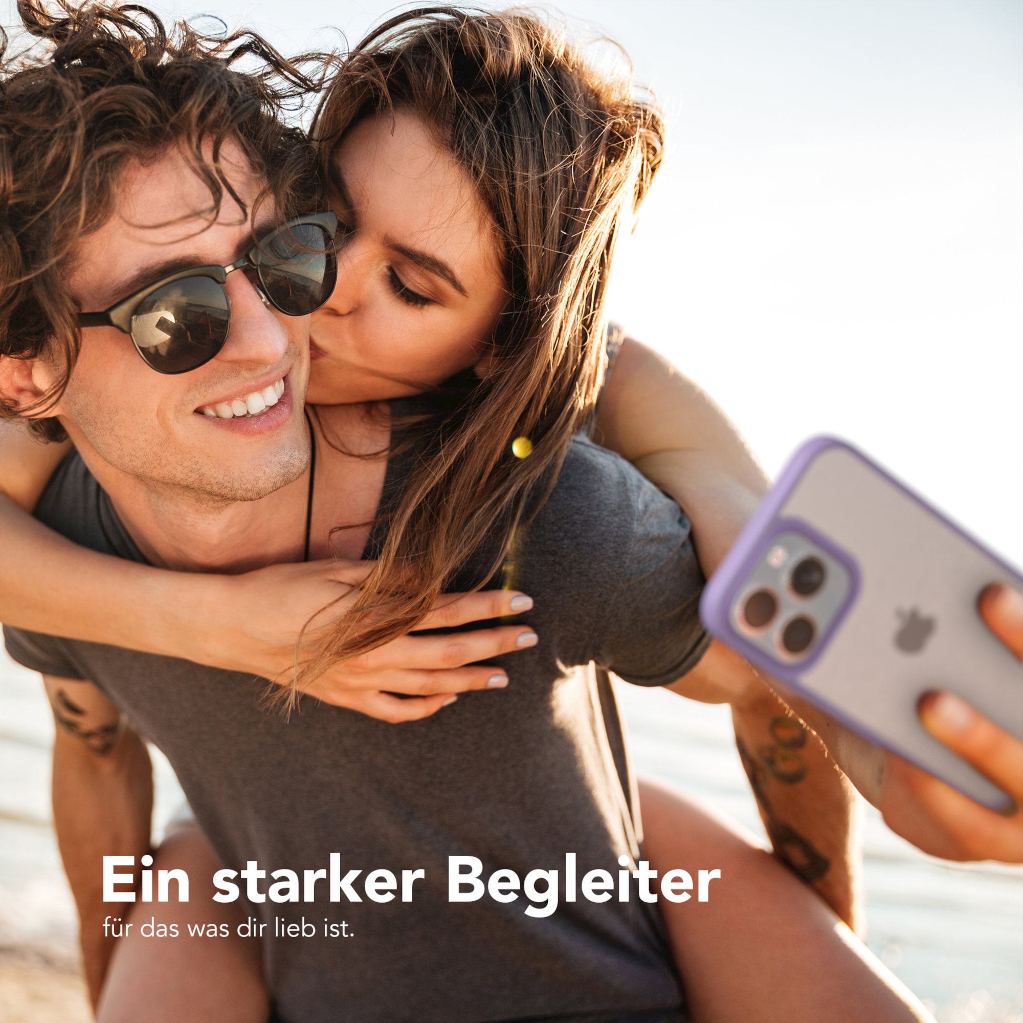 Paar macht Selfie. Frau küsst Mann. Handyhülle sichtbar. Strand und Sonnenlicht.