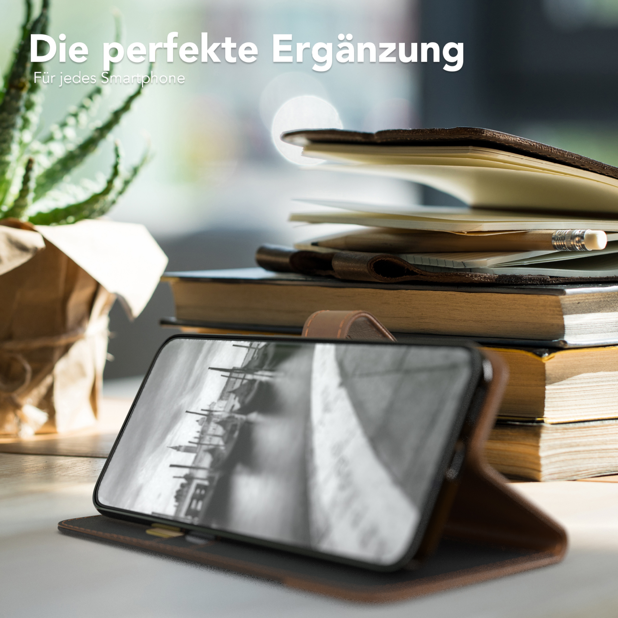 Ein Telefon in einer braunen Lederhülle, auf einem Tisch mit Büchern, einer Pflanze und einem Bleistift.
