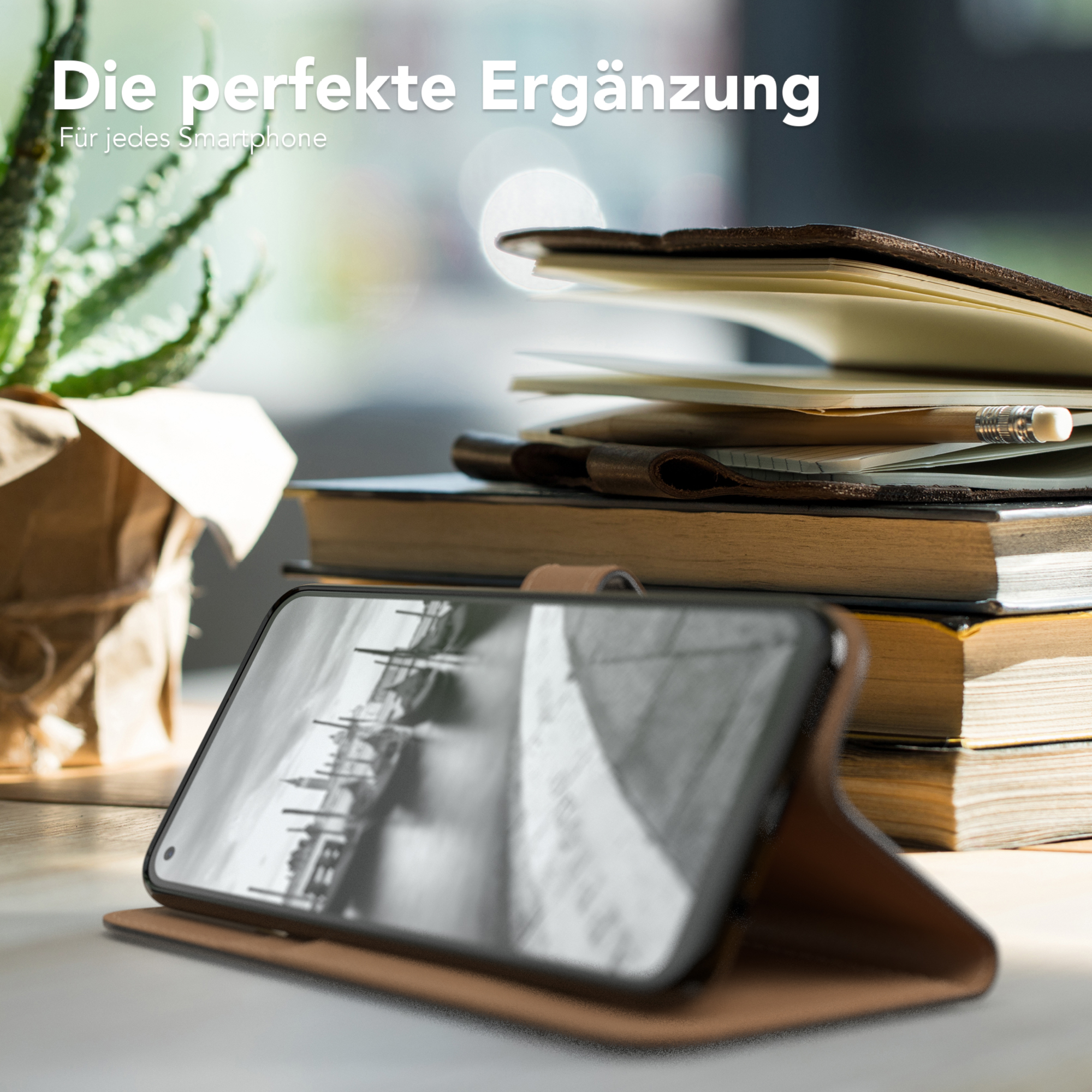 Smartphone in einem Case auf einem Tisch mit Büchern. Links ist eine Pflanze. Der Hintergrund ist verschwommen.