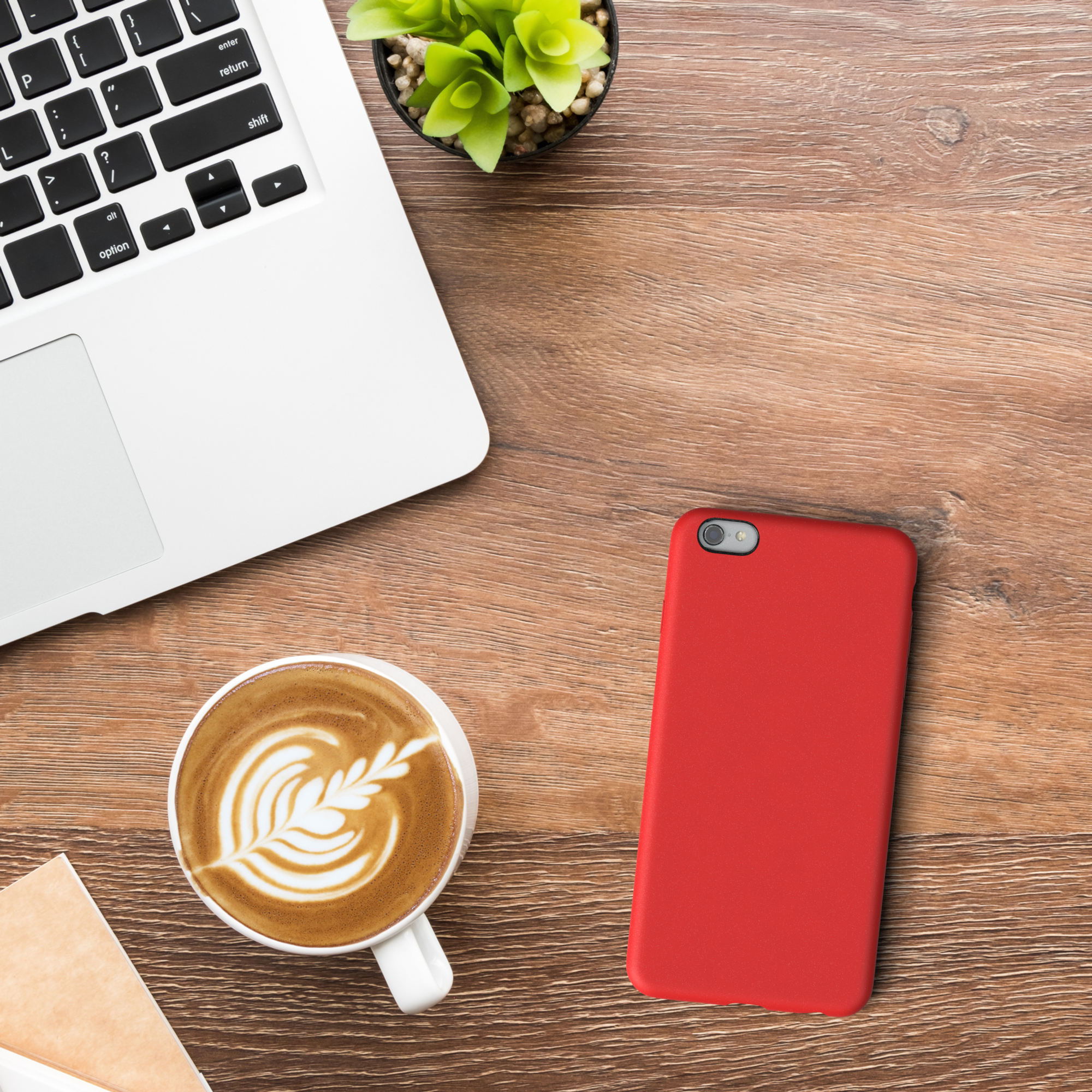 Ein Laptop, eine Tasse Kaffee mit Latte Art, eine kleine Pflanze und eine rote Handyhülle auf einem Holzschreibtisch.