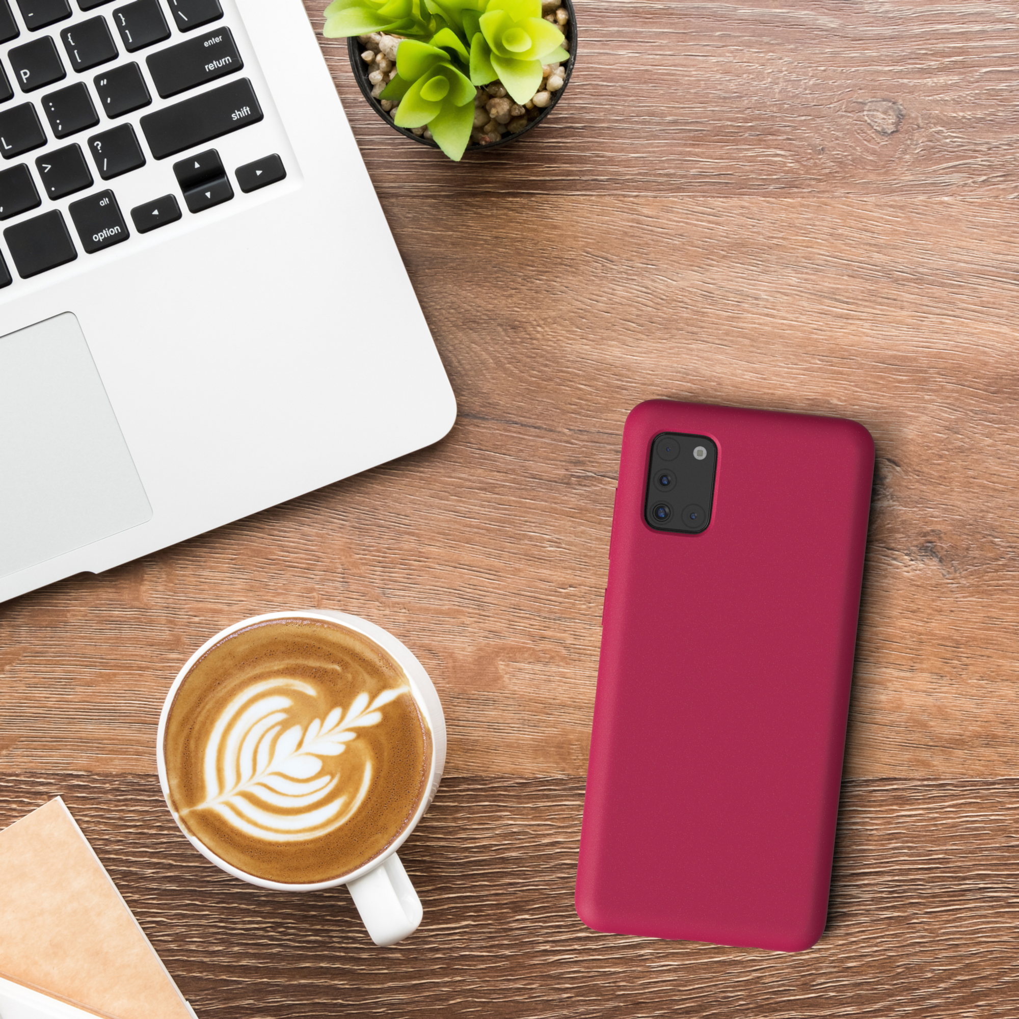 Ein Laptop, eine Kaffeetasse mit Kunst, eine rote Handyhülle und eine kleine Pflanze auf einem Holztisch.