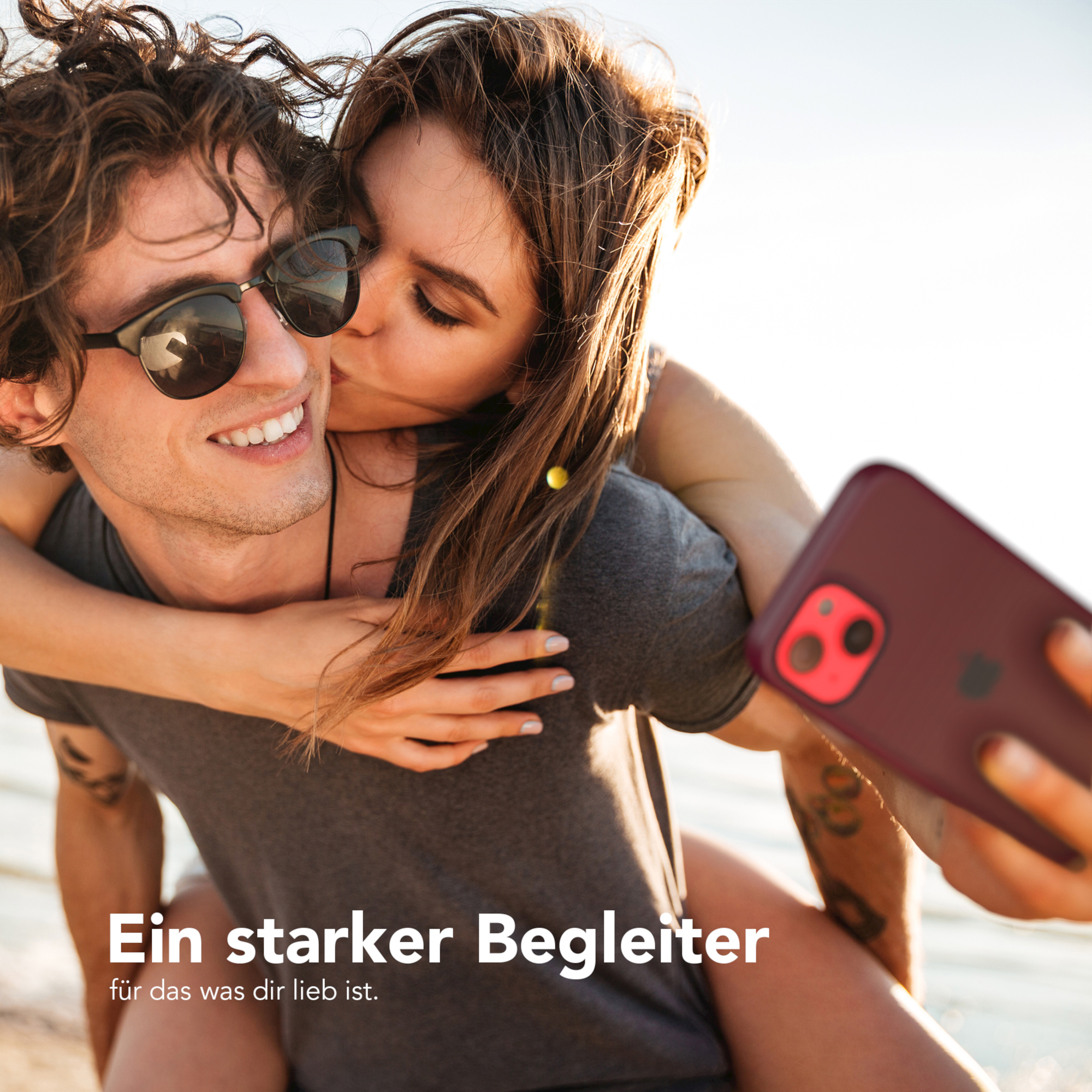 Ein Mann und eine Frau küssen sich. Die Frau trägt eine Sonnenbrille und hält ein Telefon. Der Hintergrund ist verschwommen.