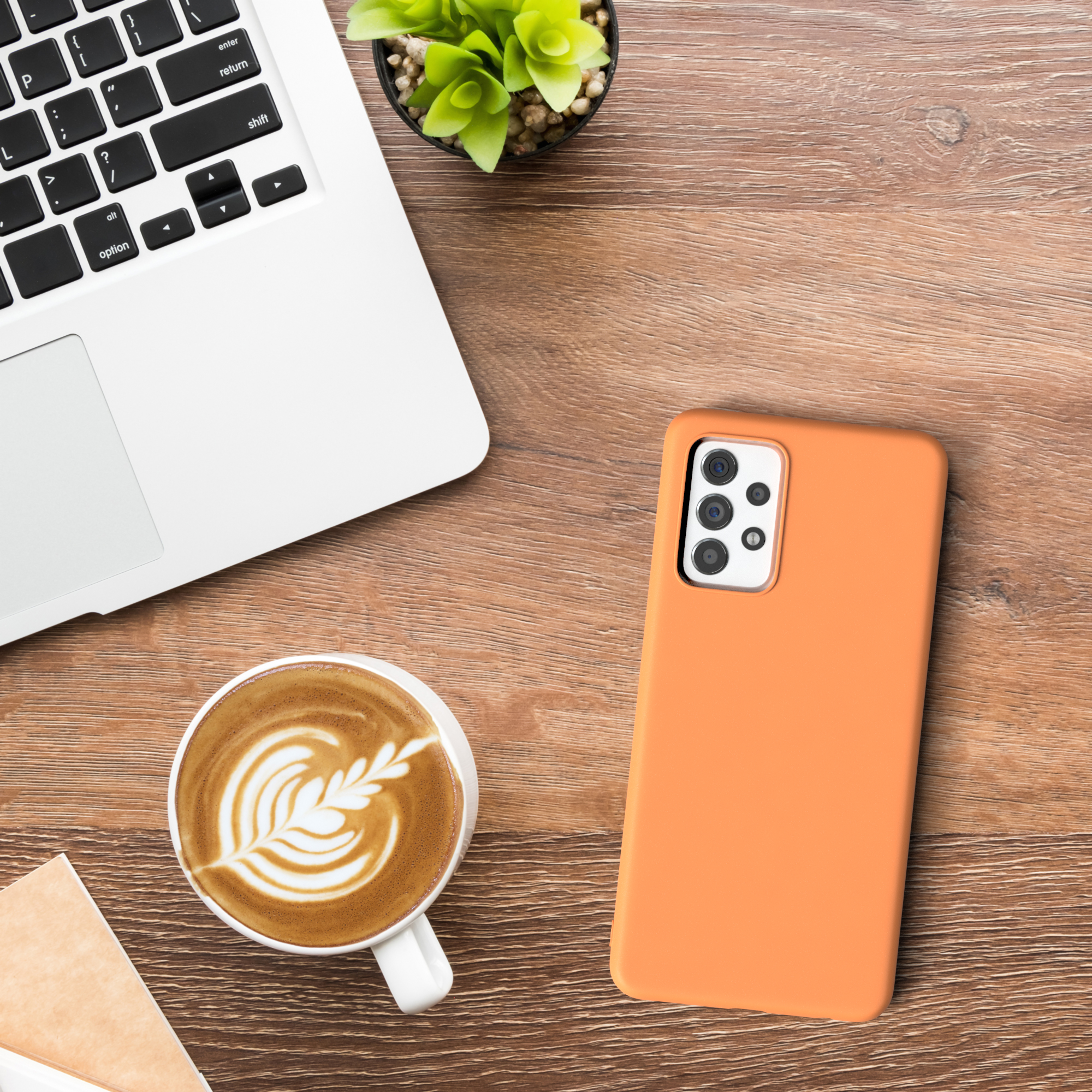 Orangefarbene Handyhülle, Laptop, Kaffeetasse mit Design auf einem Holzschreibtisch. Grüne Pflanze.