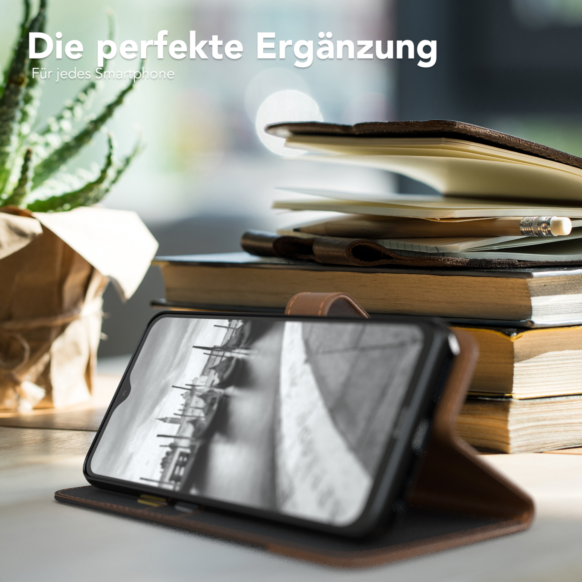 Ein Telefon in einer Hülle liegt auf einem Tisch, mit einem Buch, einem Notizbuch und einer Pflanze in der Nähe.