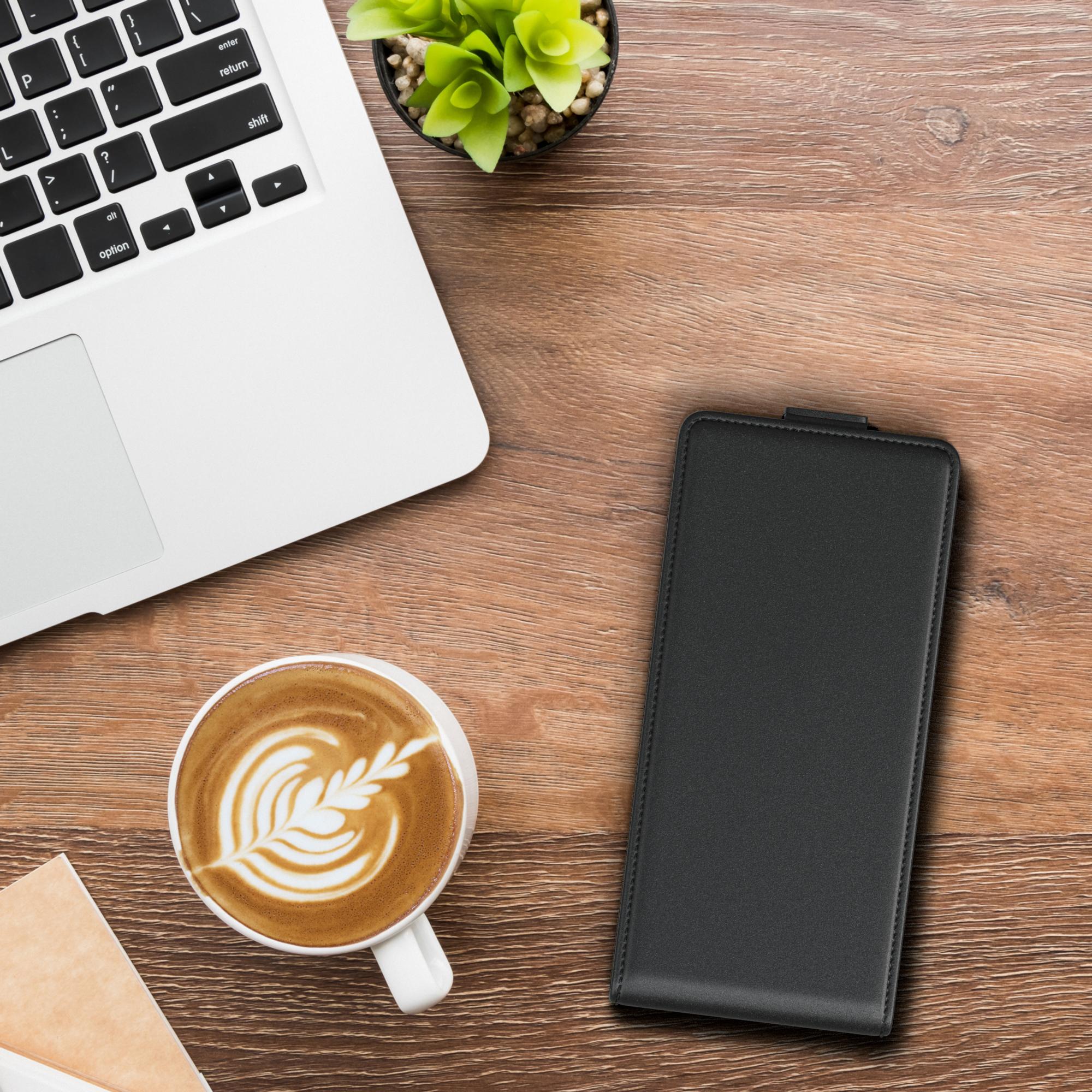 Ein Laptop, eine Tasse Kaffee, eine Pflanze und eine schwarze Handyhülle auf einem Holztisch.