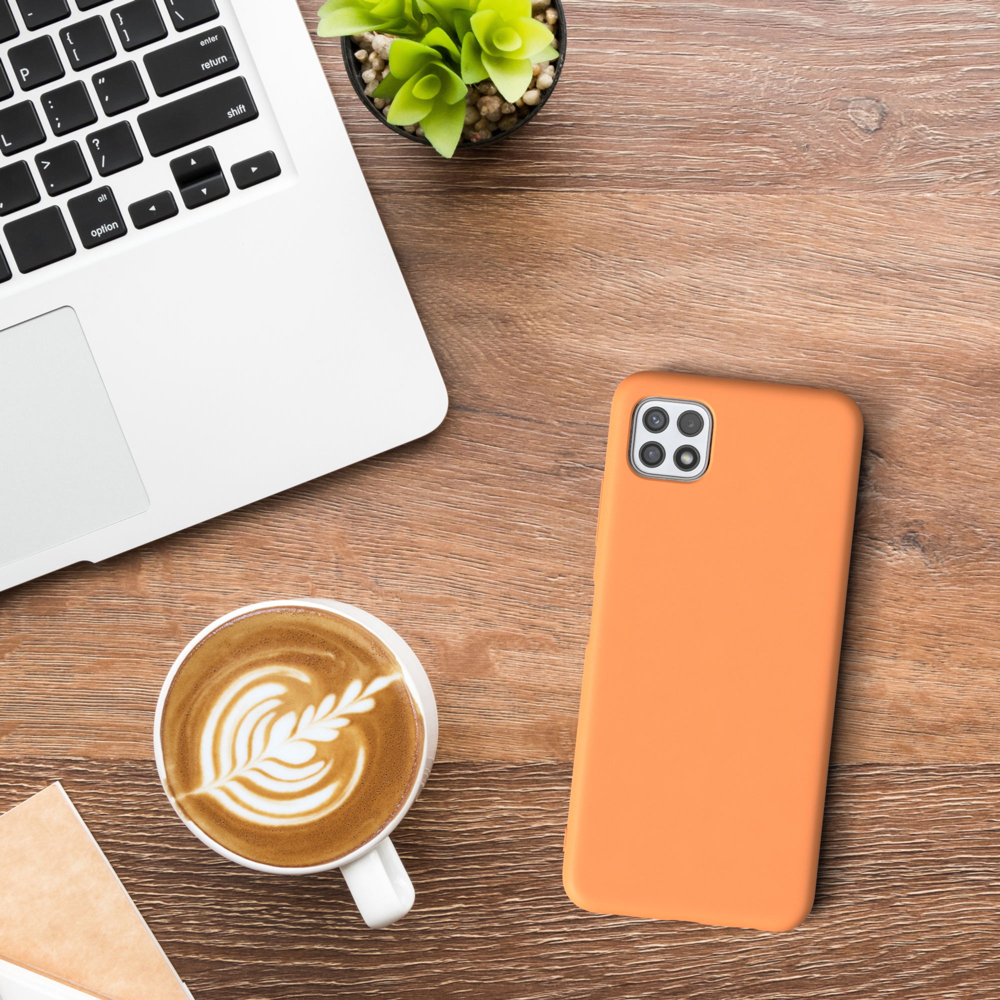 Eine orangefarbene Handyhülle liegt auf einem Holztisch. Ein Laptop, Kaffee und eine Pflanze stehen auf dem Tisch.