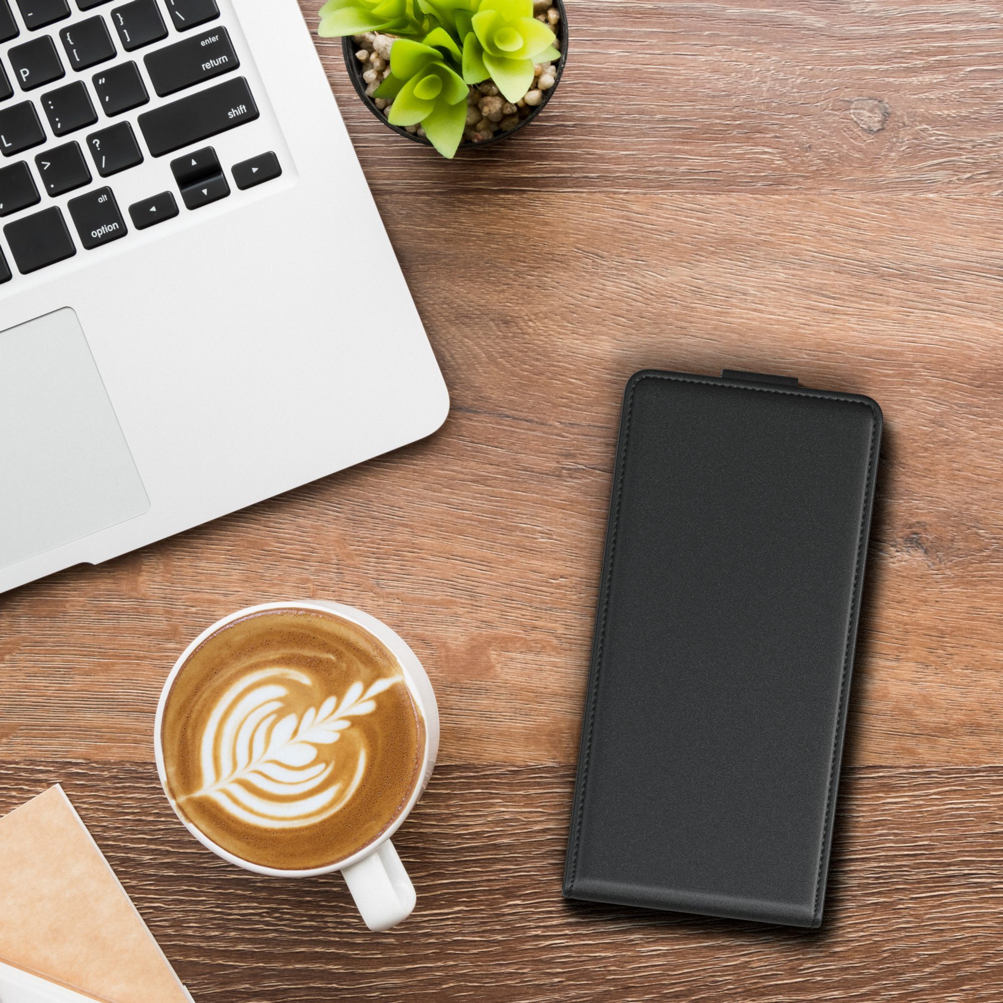Ein Laptop, eine Kaffeetasse mit Latte Art, eine grüne Pflanze und eine geschlossene schwarze Handyhülle auf einem Holztisch.