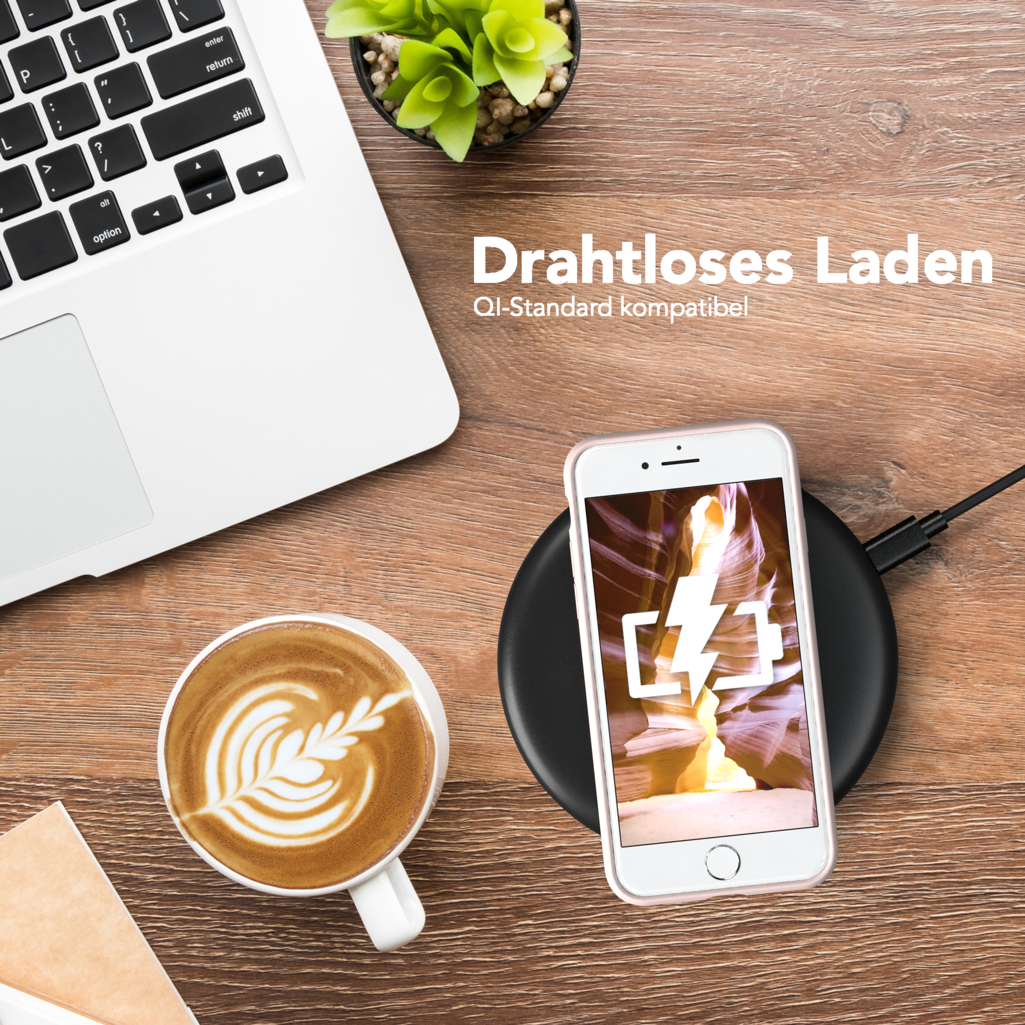 Ein Laptop, eine Kaffeetasse mit Latte Art, ein Telefon auf einem kabellosen Ladegerät und eine kleine Pflanze.