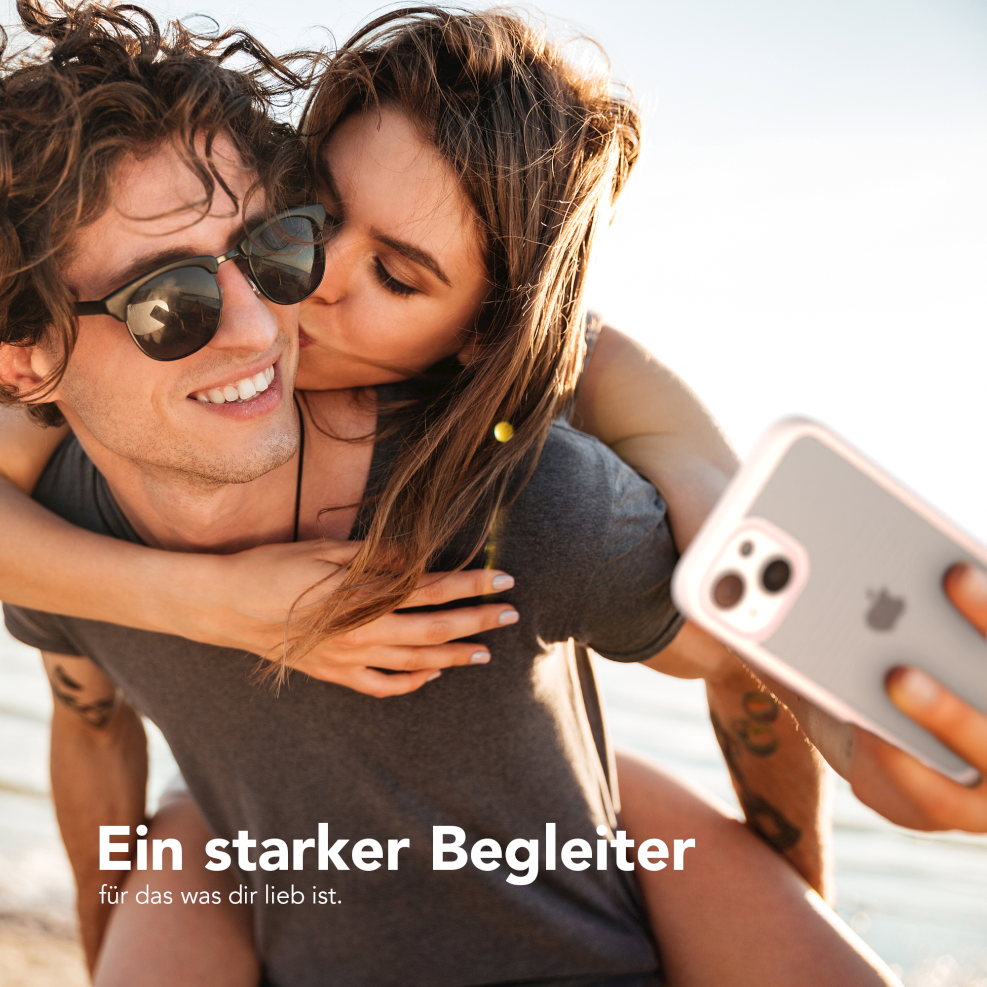 Ein Paar, Frau küsst Mann, hält Telefon mit klarer Hülle. Strandhintergrund.