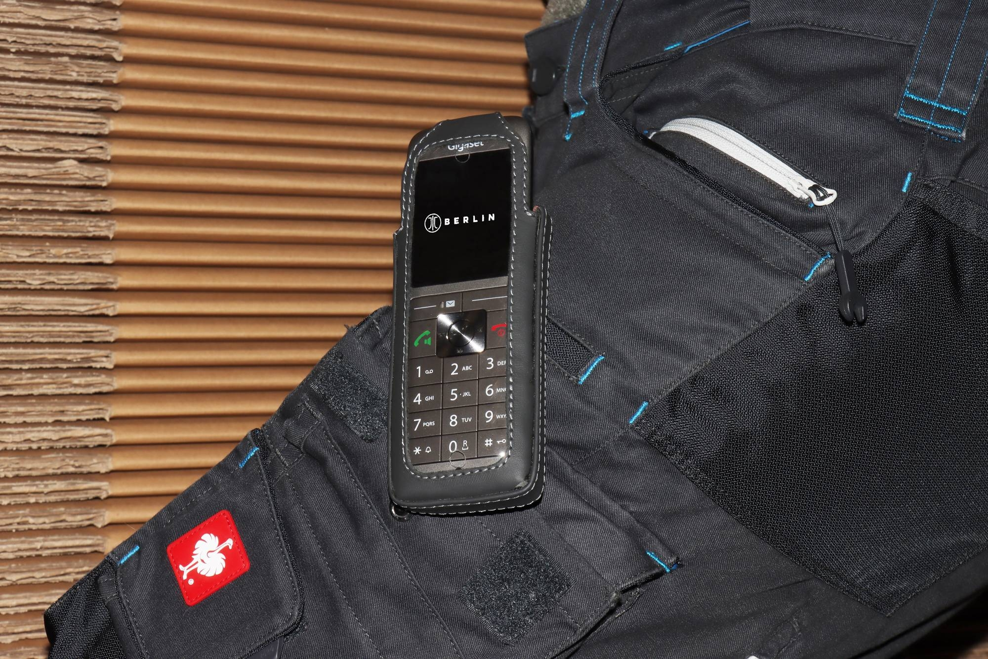 Ein Telefon in einer Hülle auf einem Arbeitshosenbein, mit rotem Logo und Nähten.