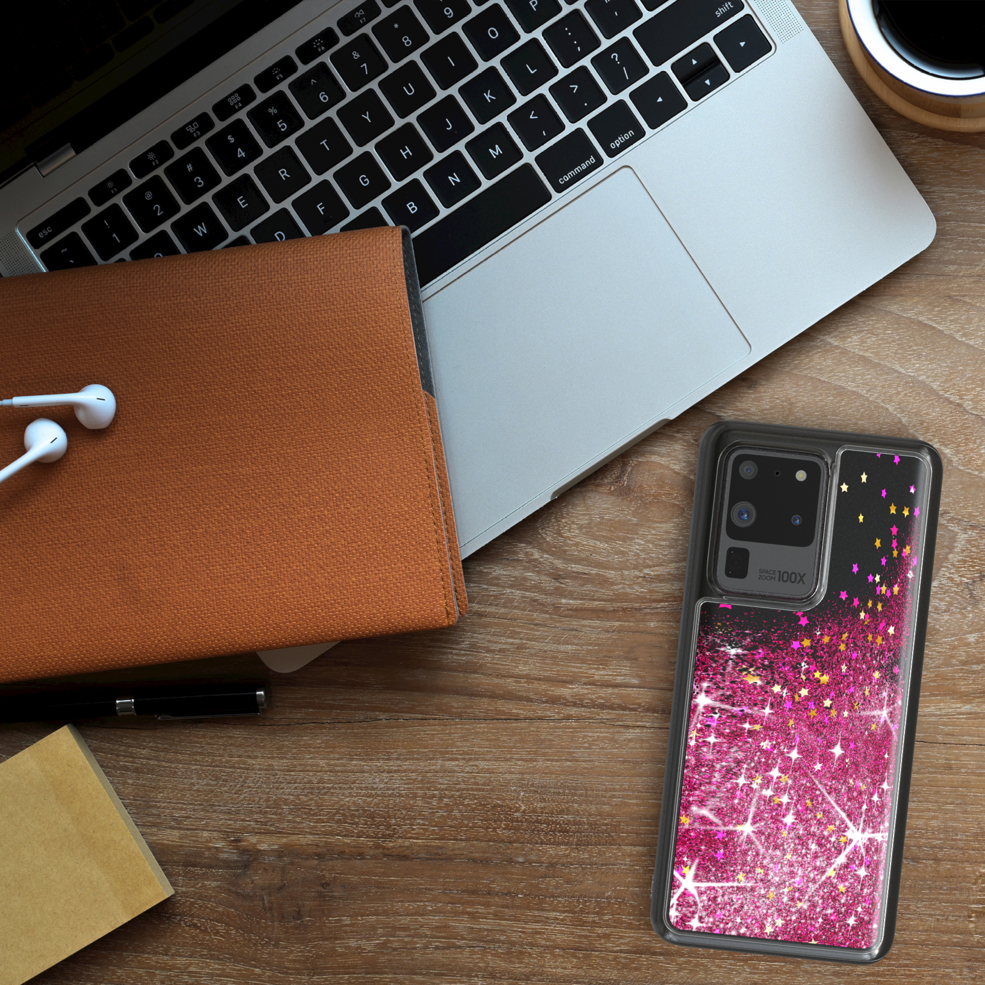 Laptop, Notizbuch, Handyhülle und Ohrhörer auf einem Holzschreibtisch. Handyhülle hat pinkes Glitzerdesign.