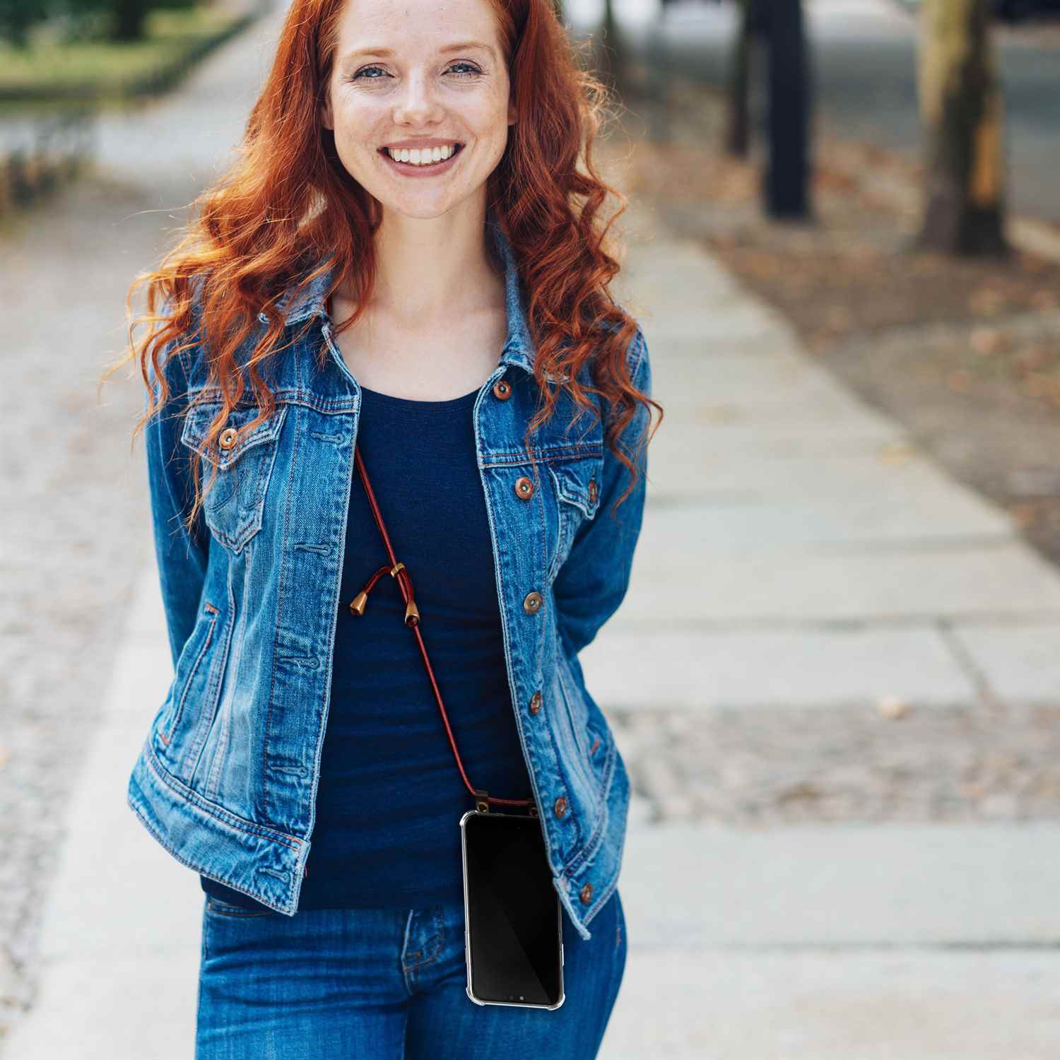 Eine Frau mit roten Haaren trägt eine Jeansjacke und trägt ein Telefon mit braunem Riemen.