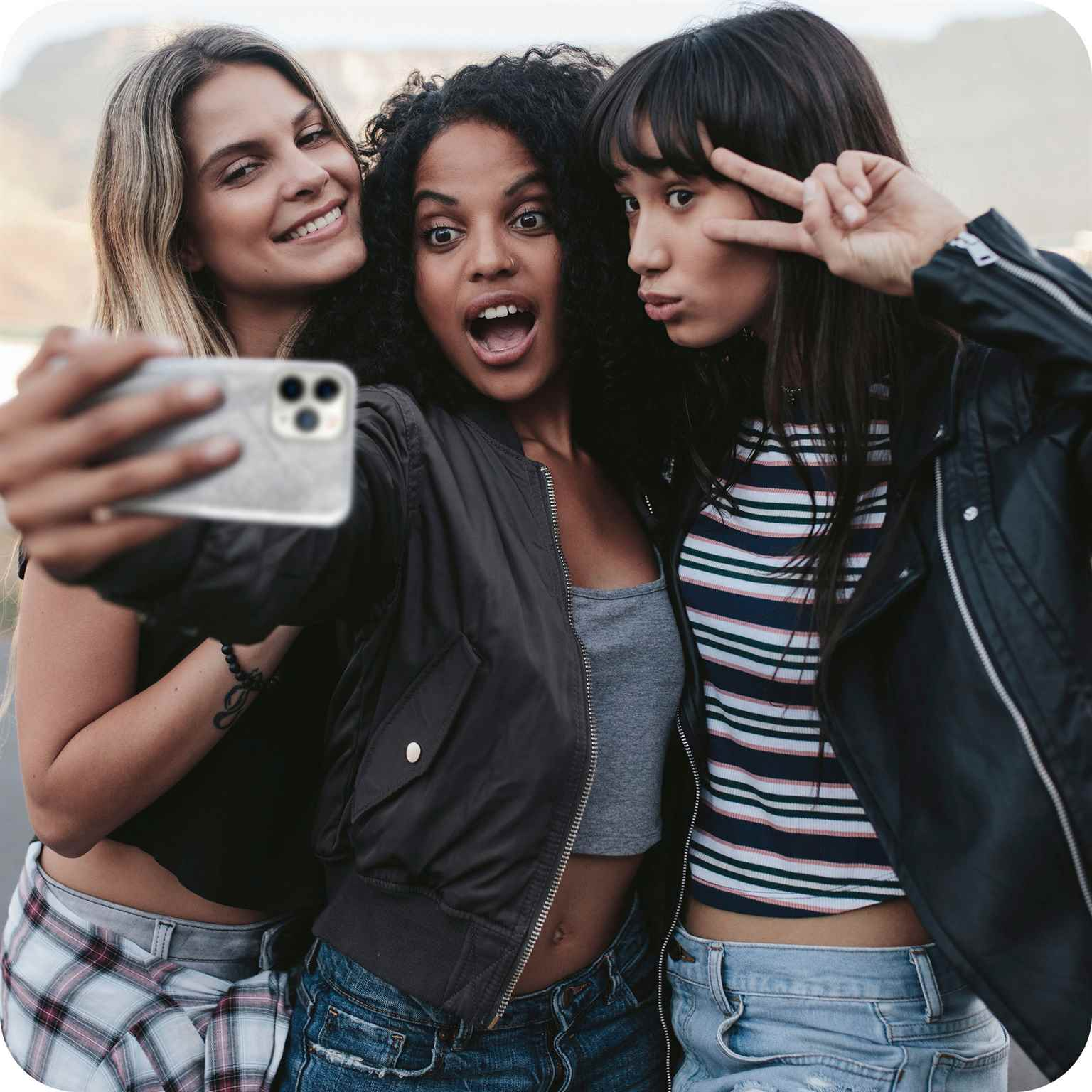 Drei Frauen machen ein Selfie mit einem Telefon. Sie lächeln und machen Gesten. Draußen.