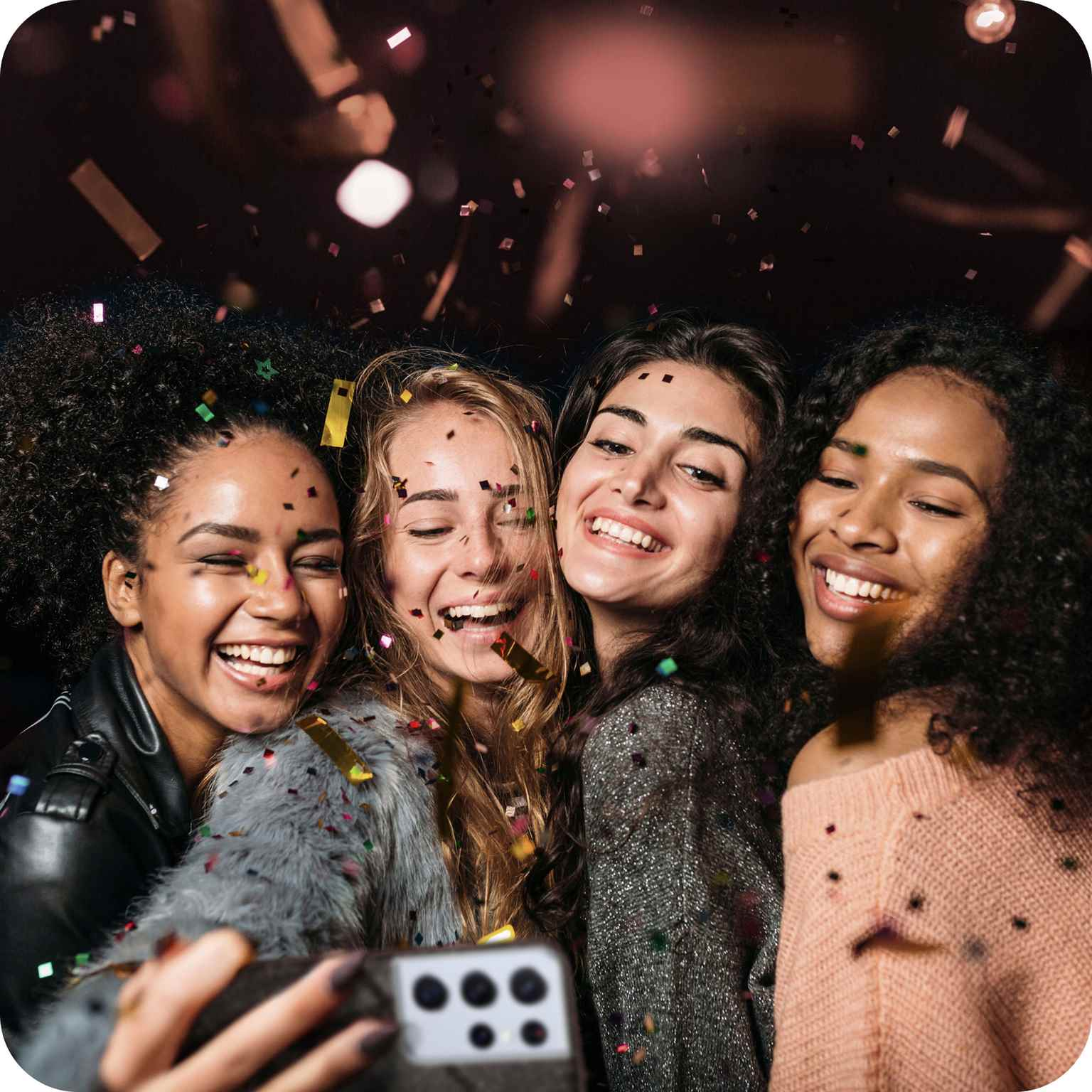 Vier Frauen lächeln, machen ein Selfie mit Konfetti in der Luft. Dunkler Hintergrund.
