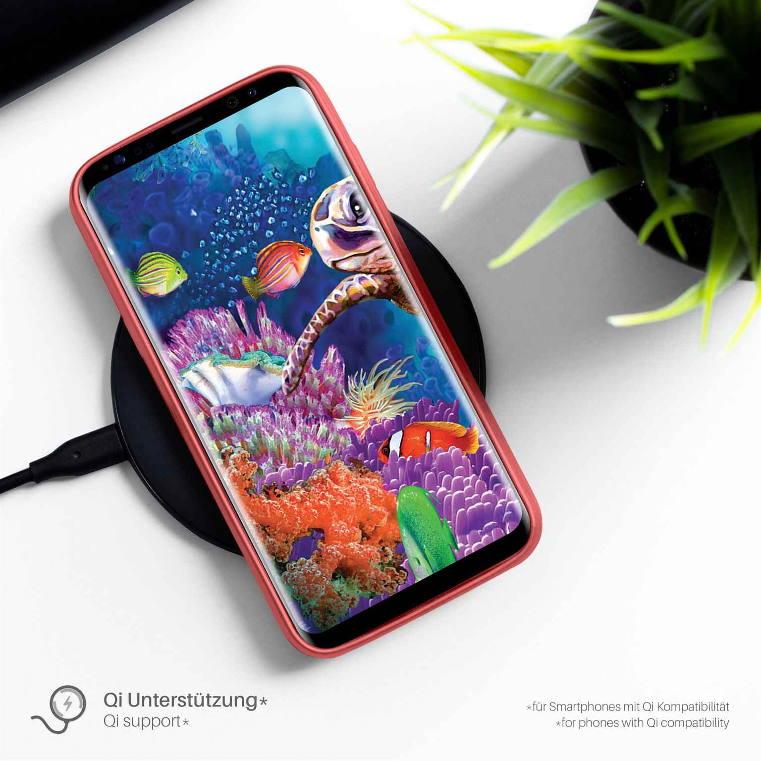 Telefon auf Ladegerät, rote Hülle, Korallen und Fische auf dem Bildschirm, Pflanze im Hintergrund.