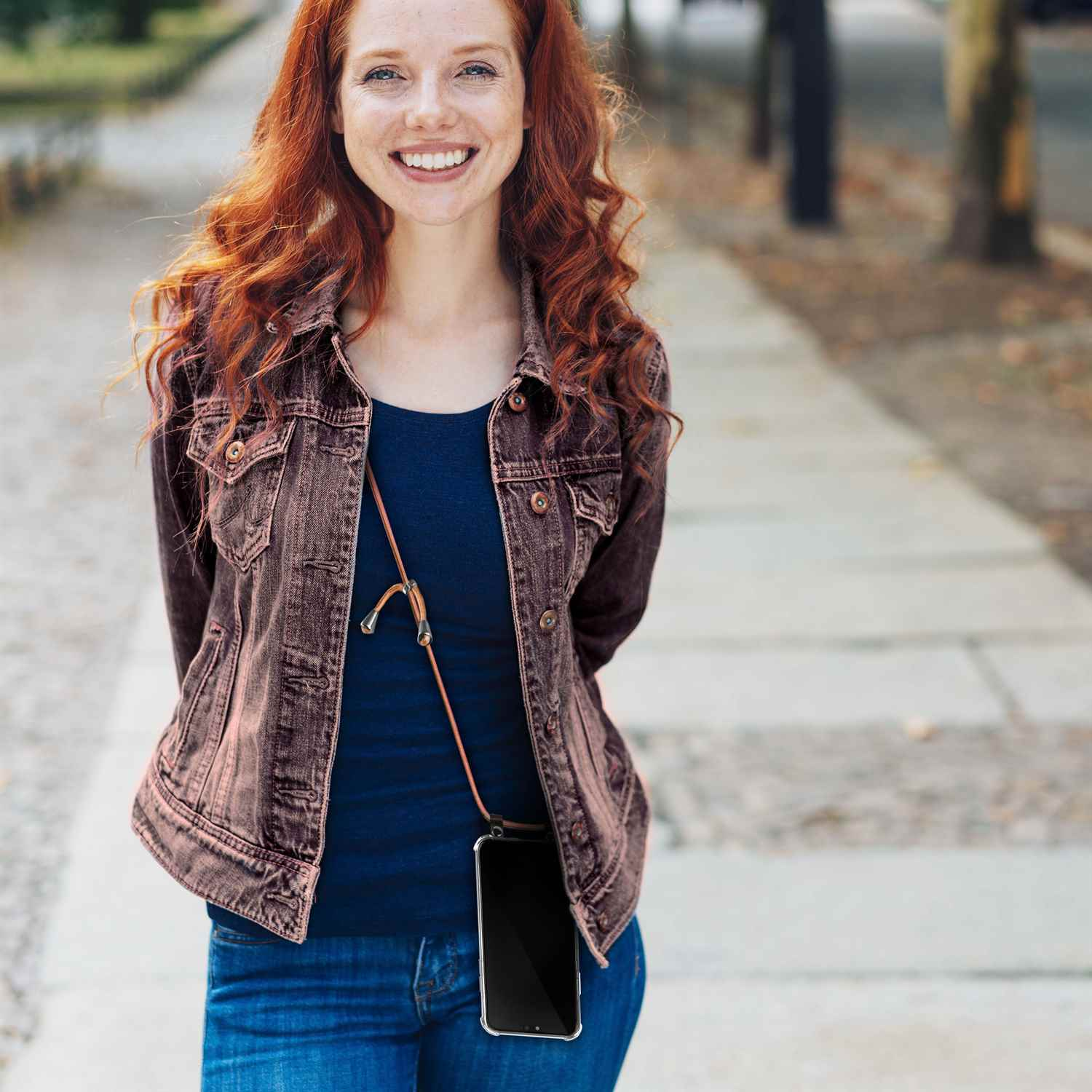 Frau mit roten Haaren trägt eine Jacke, Jeans und ein Telefon mit braunem Kordel um den Hals.