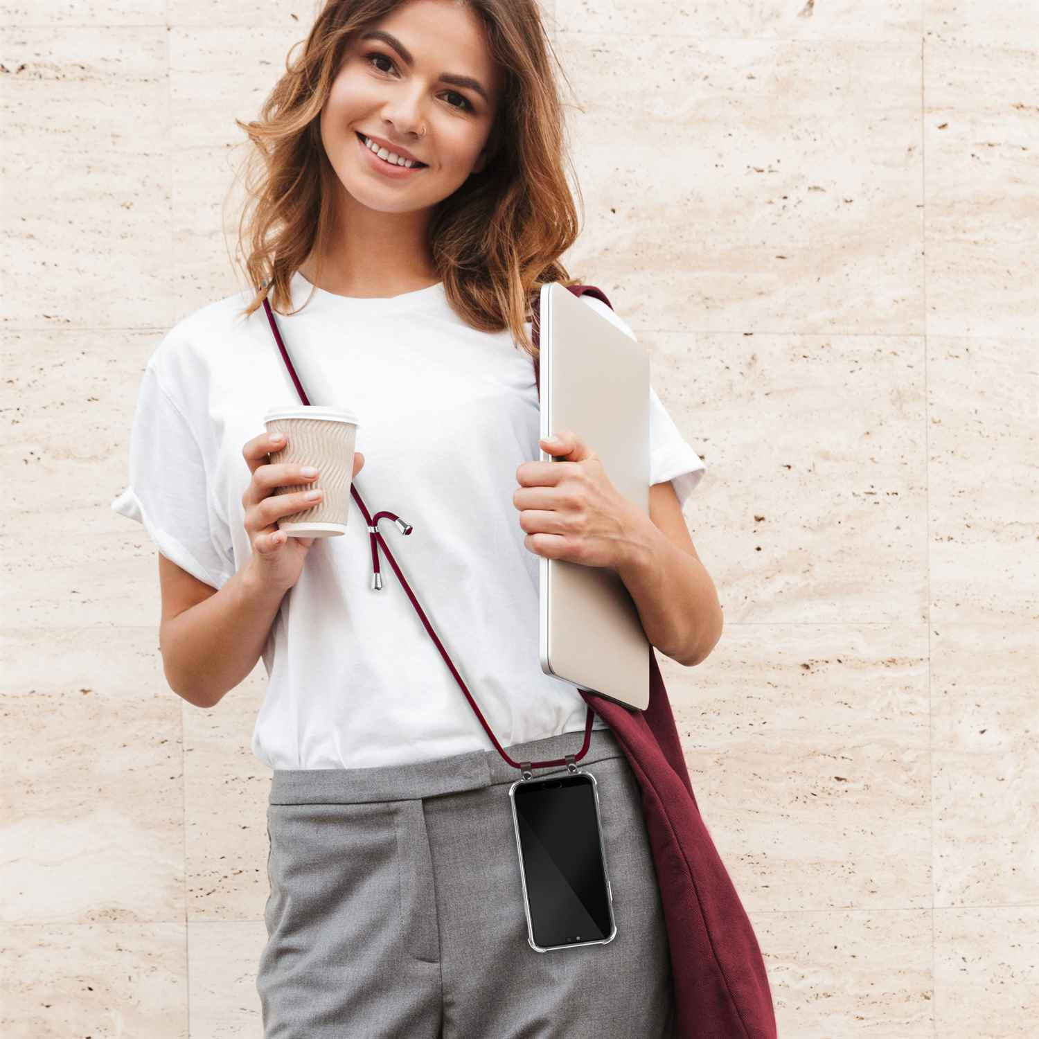 Frau mit Telefon an rotem Lanyard hält Kaffeetasse und Laptop. Beiger Wandhintergrund.