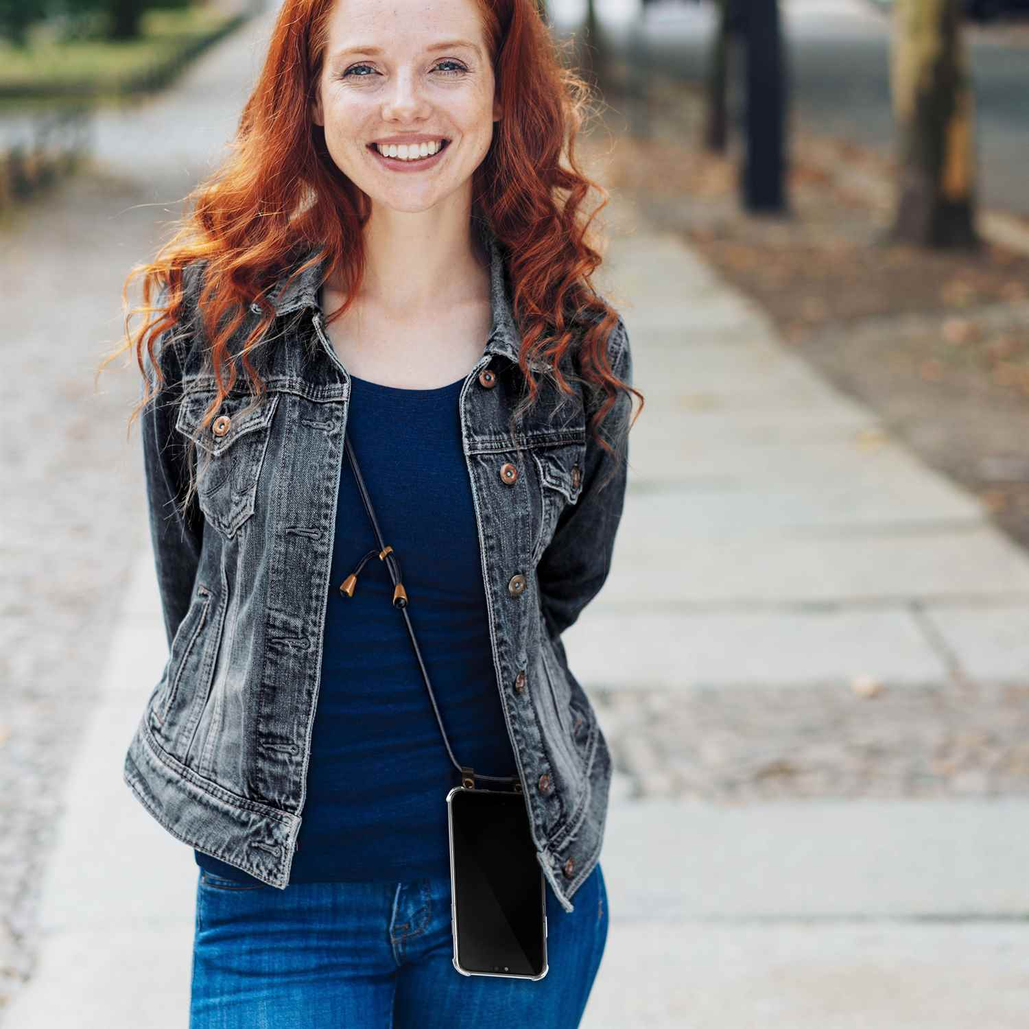Eine Frau mit roten Haaren lächelt. Sie trägt eine Jeansjacke, Jeans und ein Handy mit Lanyard.