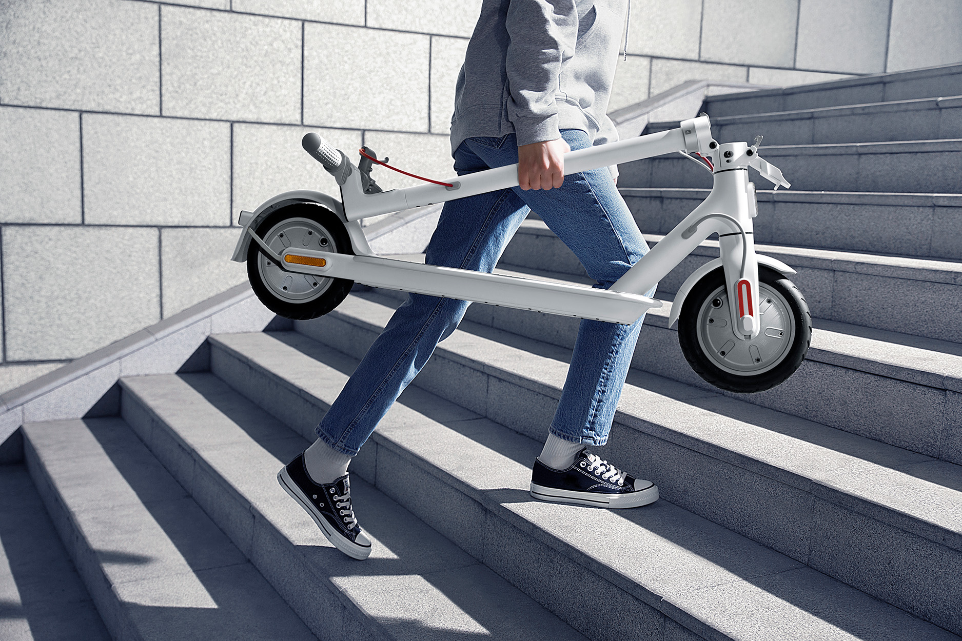 Eine Person trägt einen weißen Roller die graue Treppe hinauf. Sie trägt Jeans, Turnschuhe und einen grauen Hoodie. Der Roller ist zusammengeklappt.