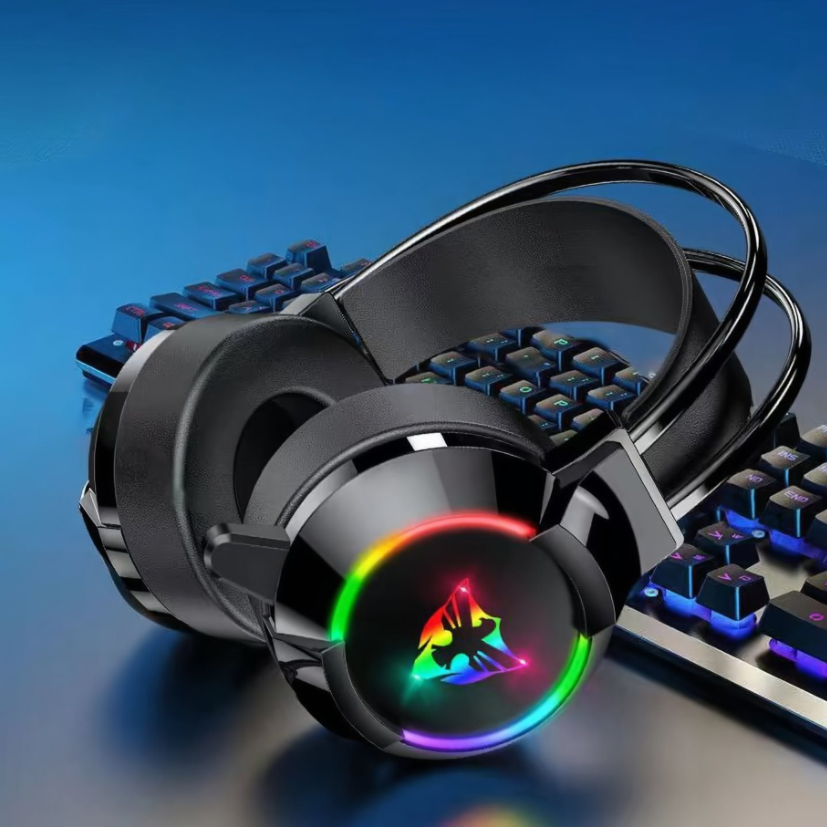 Schwarze Gaming-Kopfhörer mit Regenbogenlicht und Tastatur im Hintergrund, auf blauer Oberfläche.