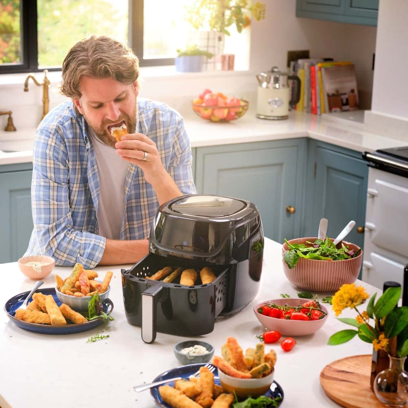 Uomo che mangia cibo fritto da un piatto. Friggitrice ad aria sul retro. Ambiente cucina con cibo sul tavolo.