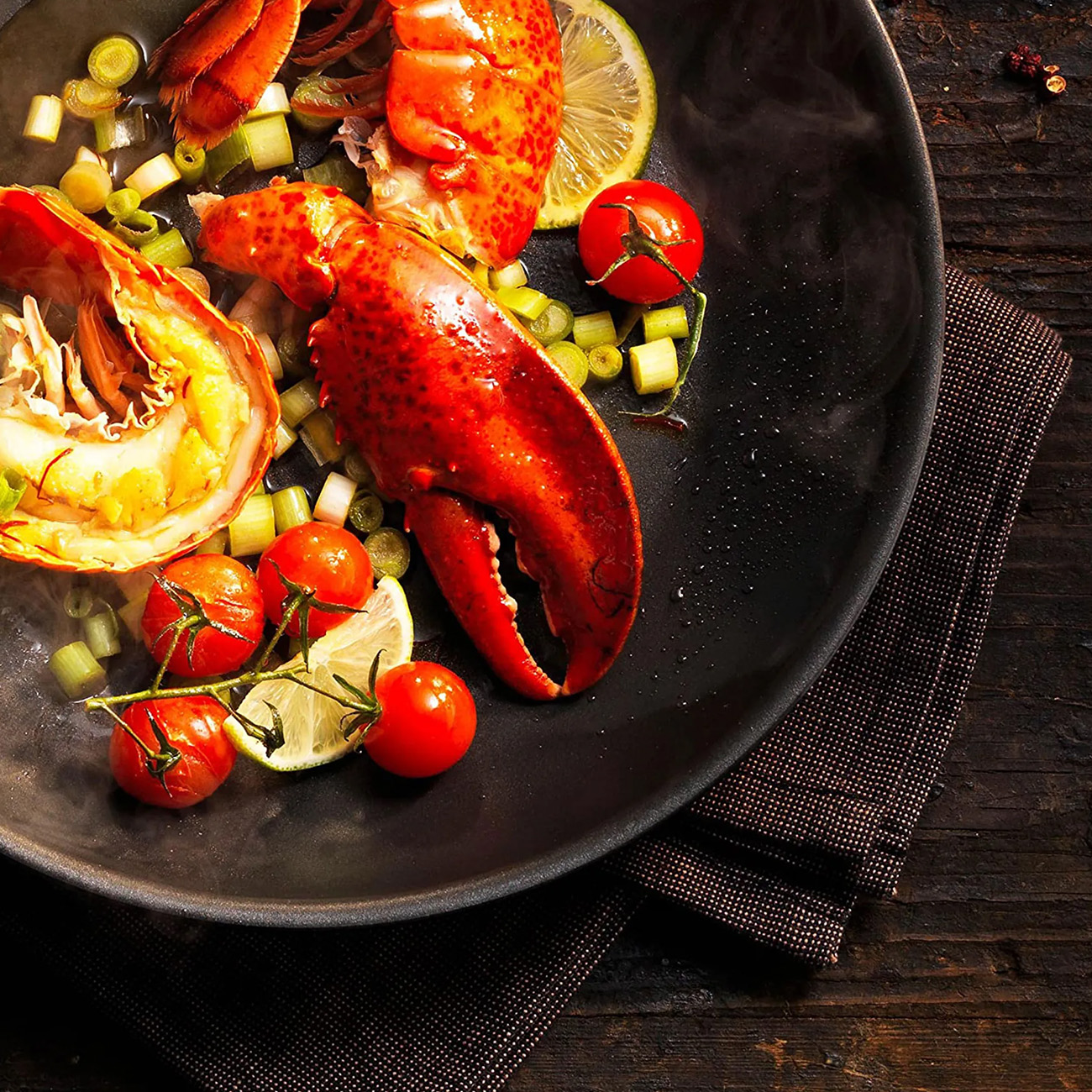 Hummergericht mit Tomaten, Zitronenscheiben und grünen Kräutern auf dunklem Teller.