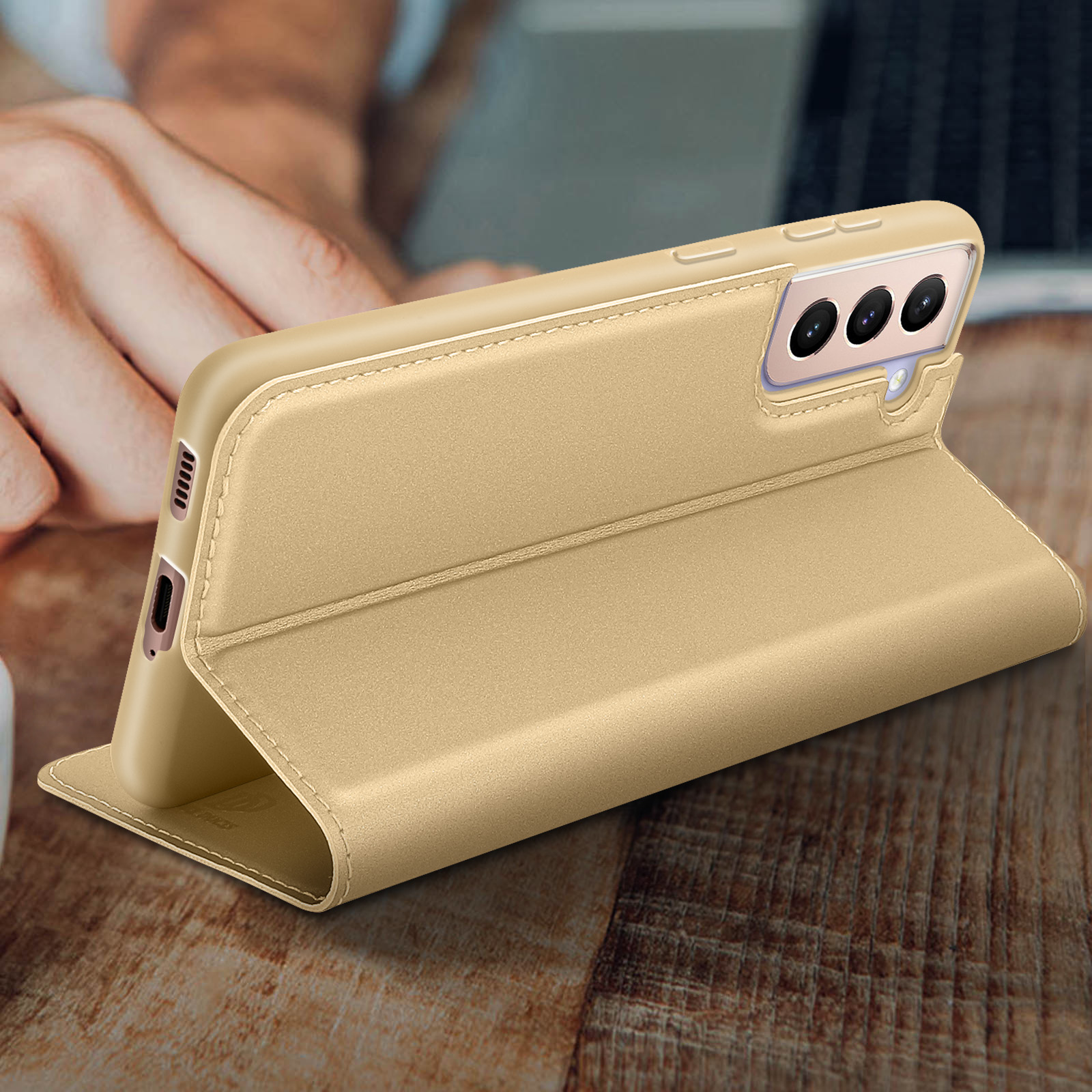 Eine goldene Handyhülle auf einem Holztisch, teilweise geöffnet. Eine Hand hält das Telefon.
