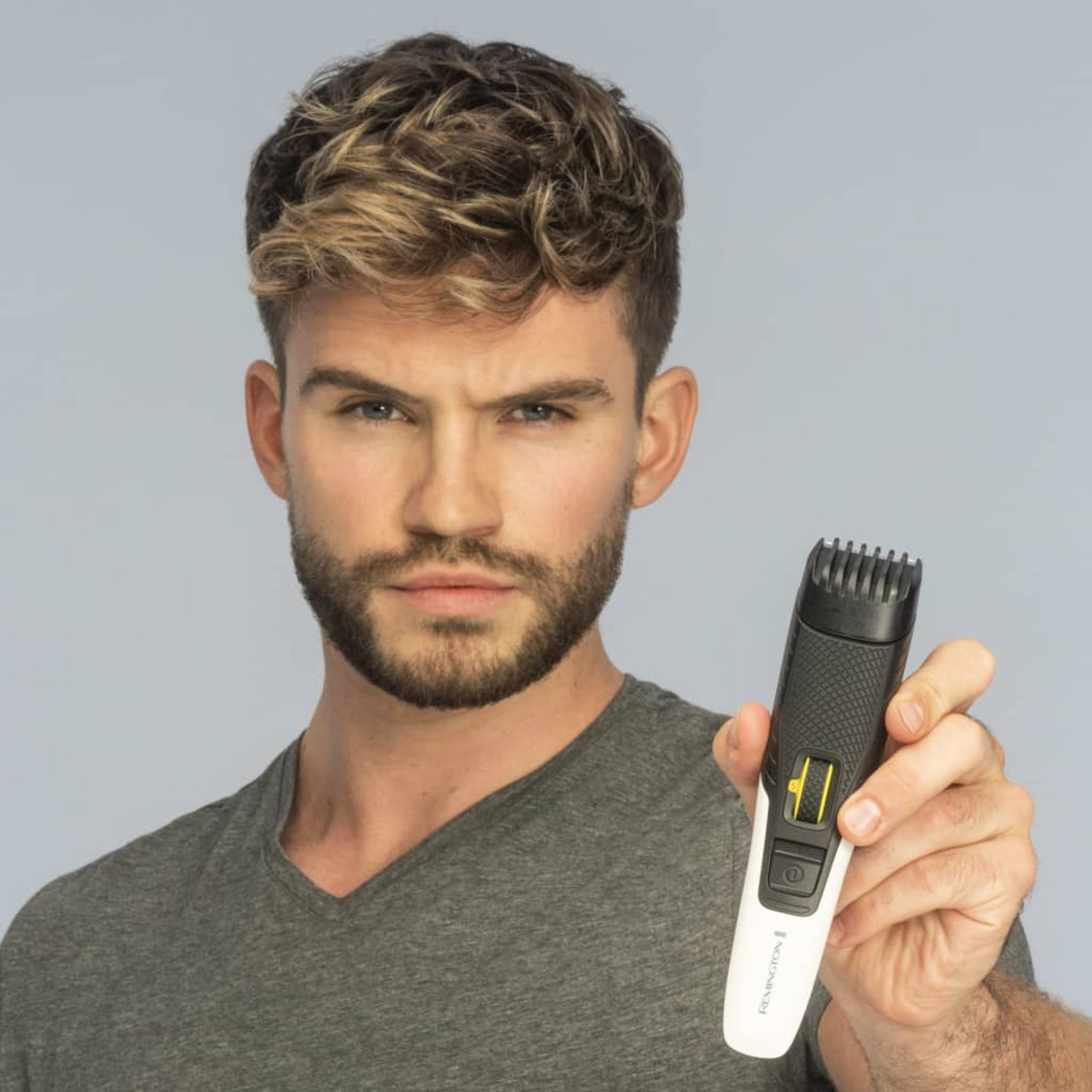 Un uomo che tiene un regolabarba, guarda la fotocamera. Ha barba e capelli biondi.
