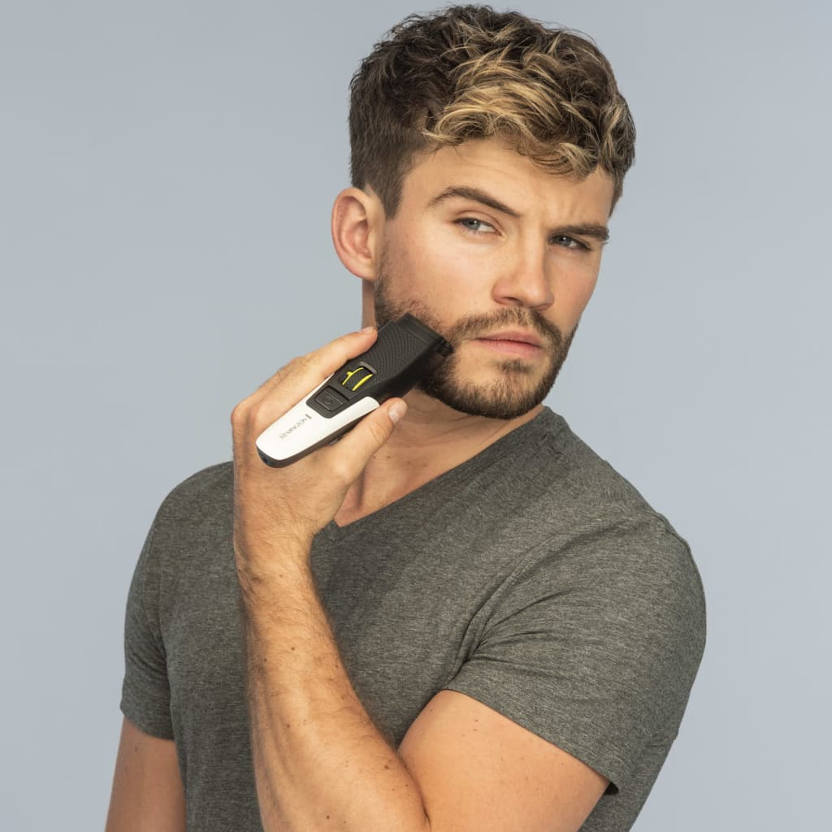 Un uomo si taglia la barba con un trimmer. Guarda la fotocamera con espressione neutra.