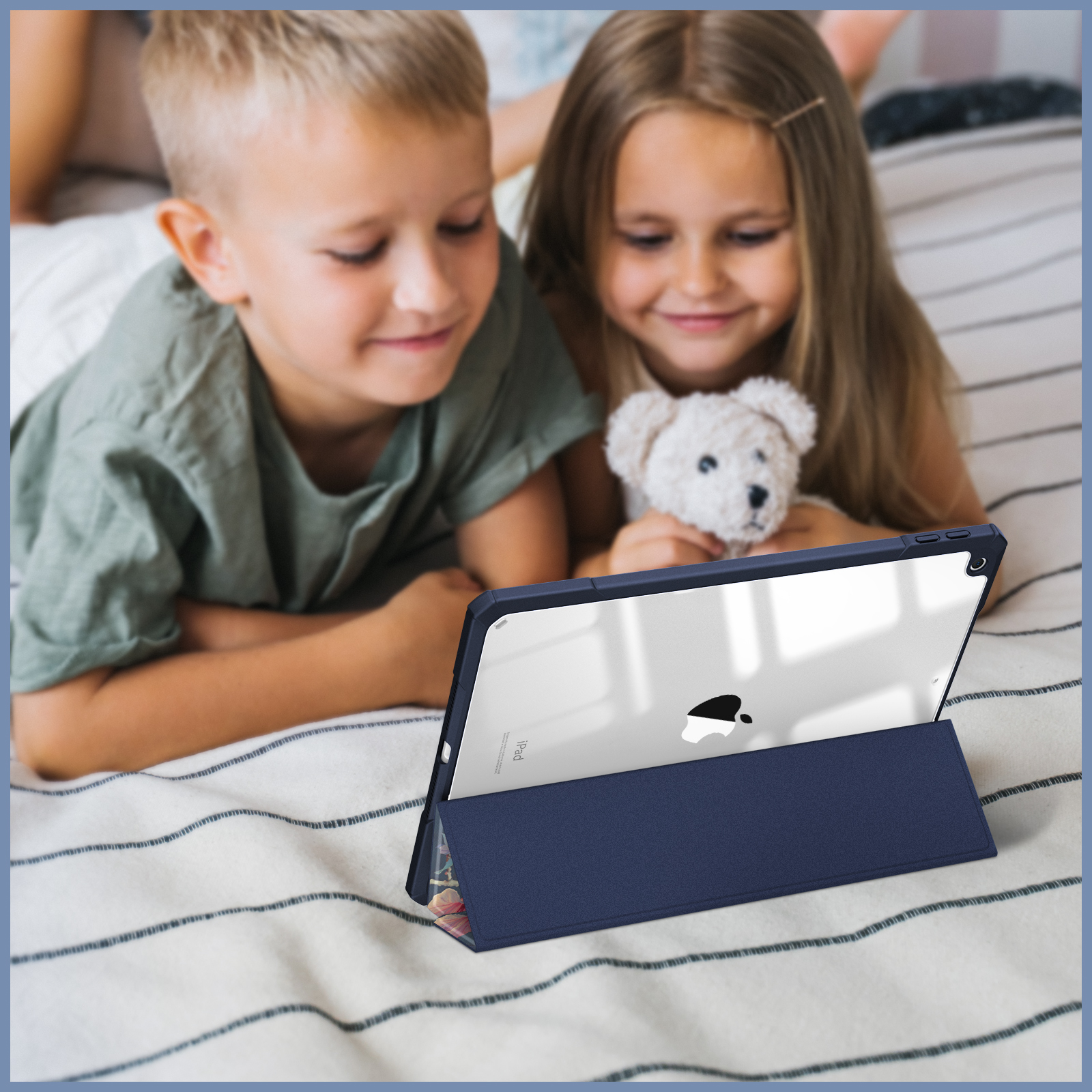 Zwei Kinder auf einem Bett mit einem Tablet in einer Hülle. Einer hält einen Teddybären.