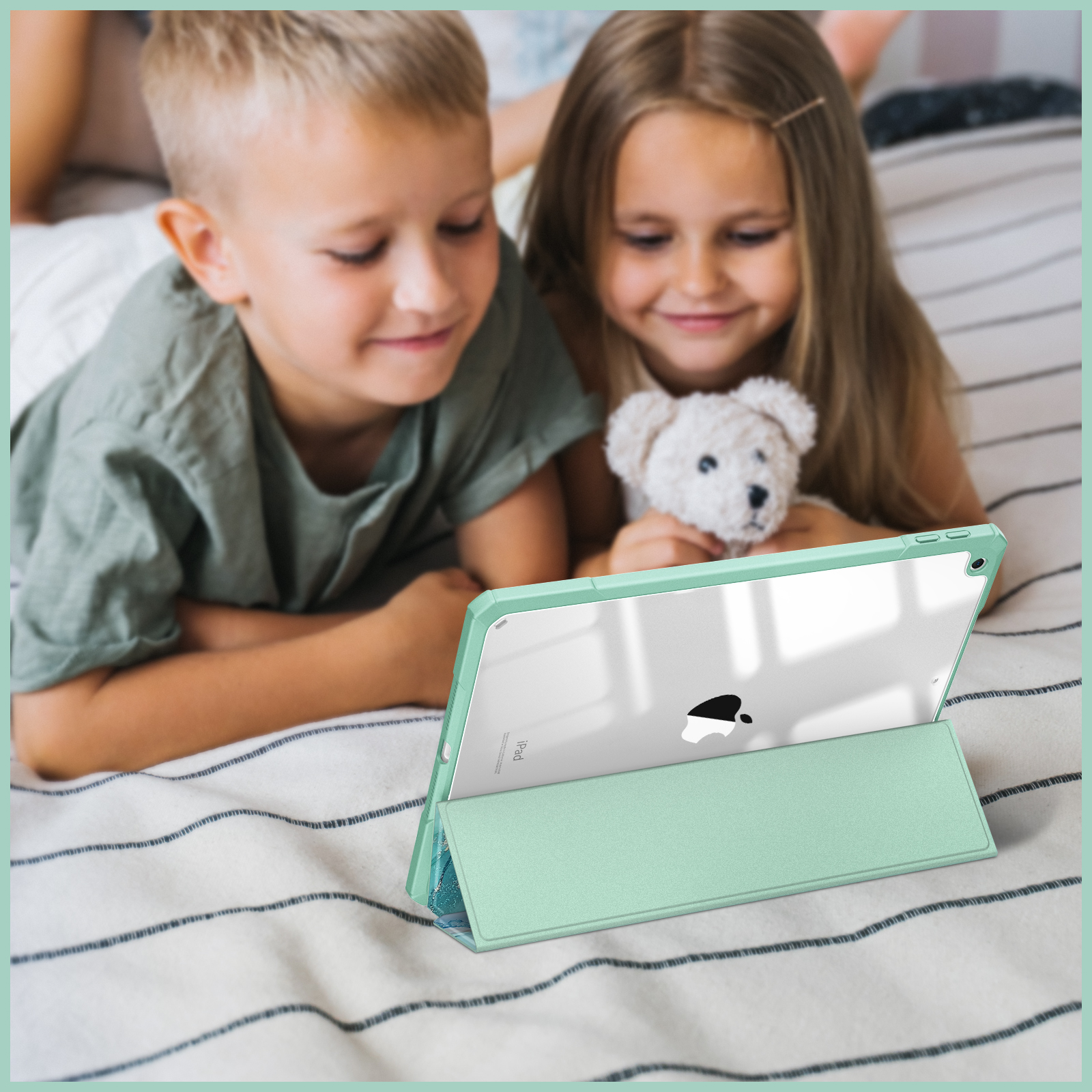 Zwei Kinder schauen auf ein Tablet. Das Tablet ist in einem grünen Case auf dem Bett.