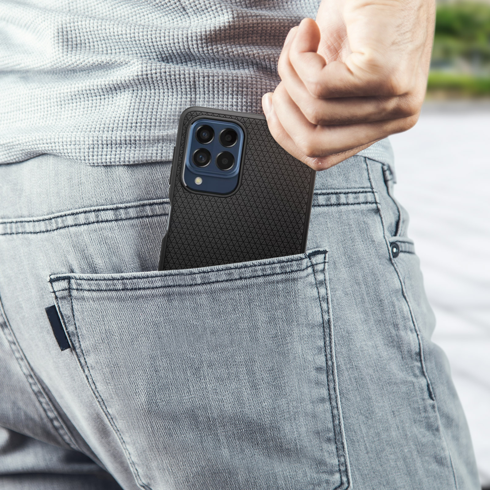 Un telefono in una custodia nera in tasca. La persona indossa una camicia grigia e jeans. Lo sfondo è sfocato.