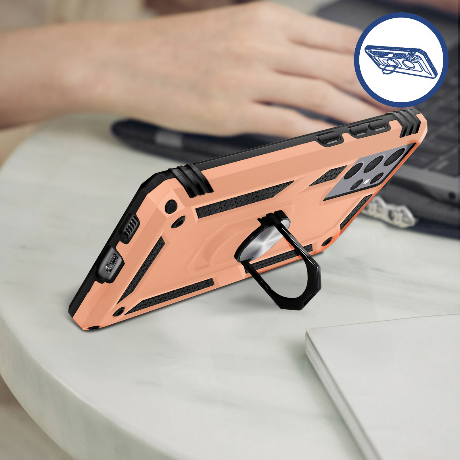 Eine rosa Handyhülle mit Ring auf einem weißen Tisch. Im Hintergrund arbeitet eine Person am Laptop.