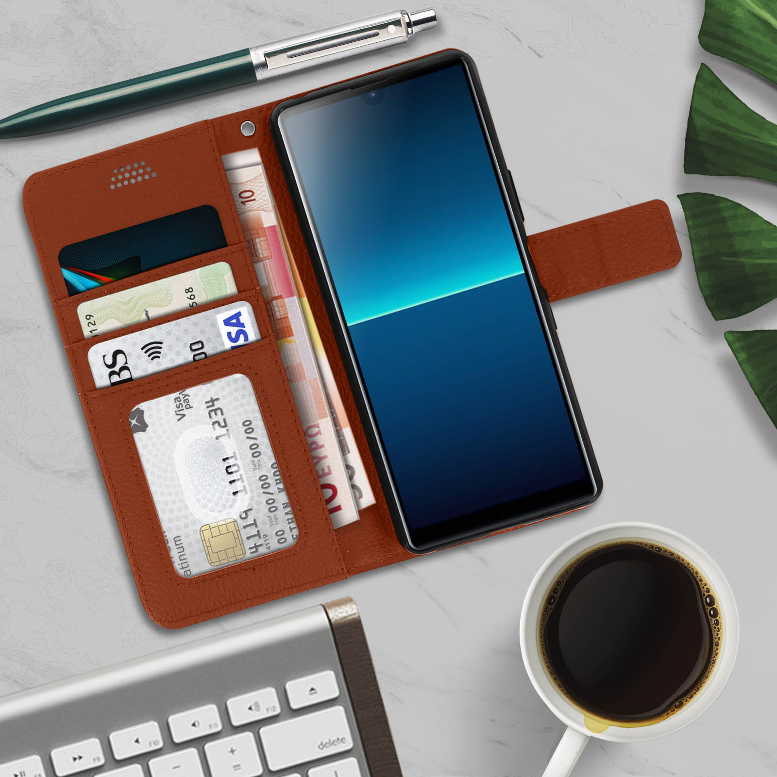 Un telefono in una custodia di pelle marrone con carte, denaro, una penna e una tazza di caffè su una superficie di marmo.