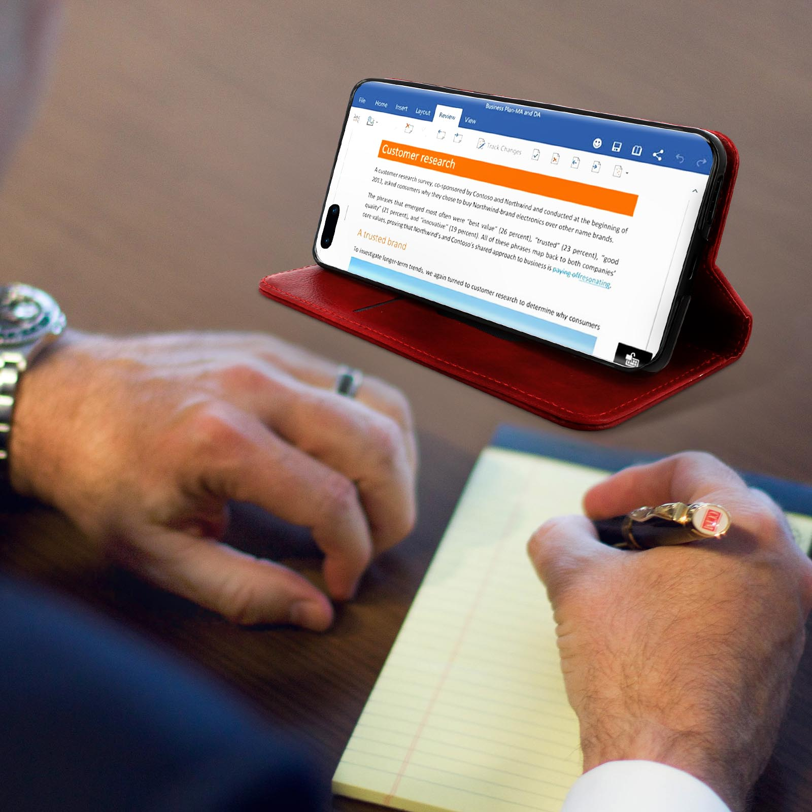 Una persona scrive in un taccuino, un telefono in una custodia rossa mostra 'Customer Research'.