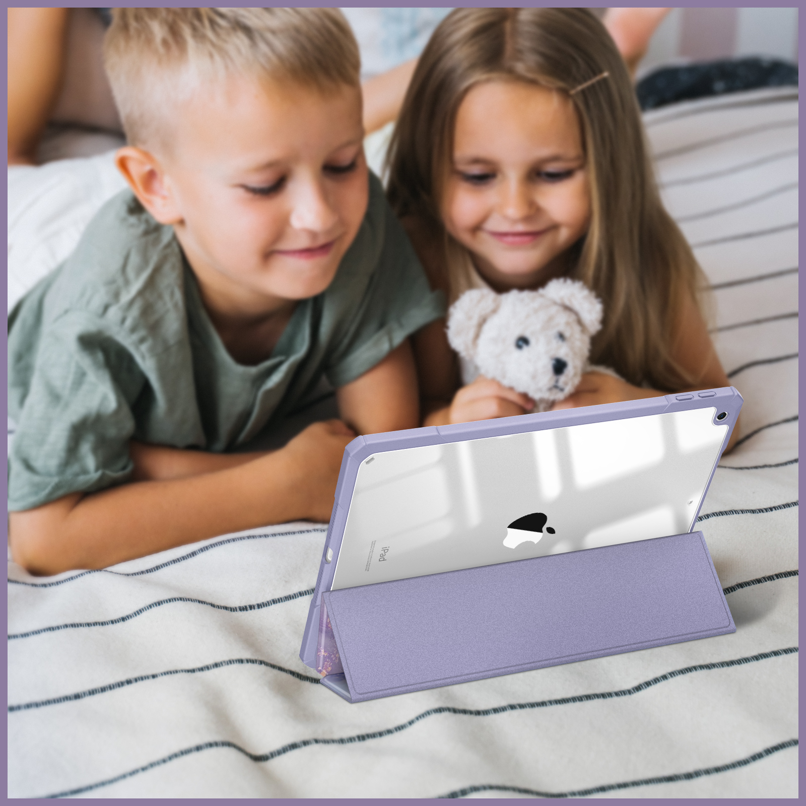 Zwei Kinder auf einem Bett schauen auf ein Tablet in einer lila Hülle. Ein Teddybär ist auch dabei.