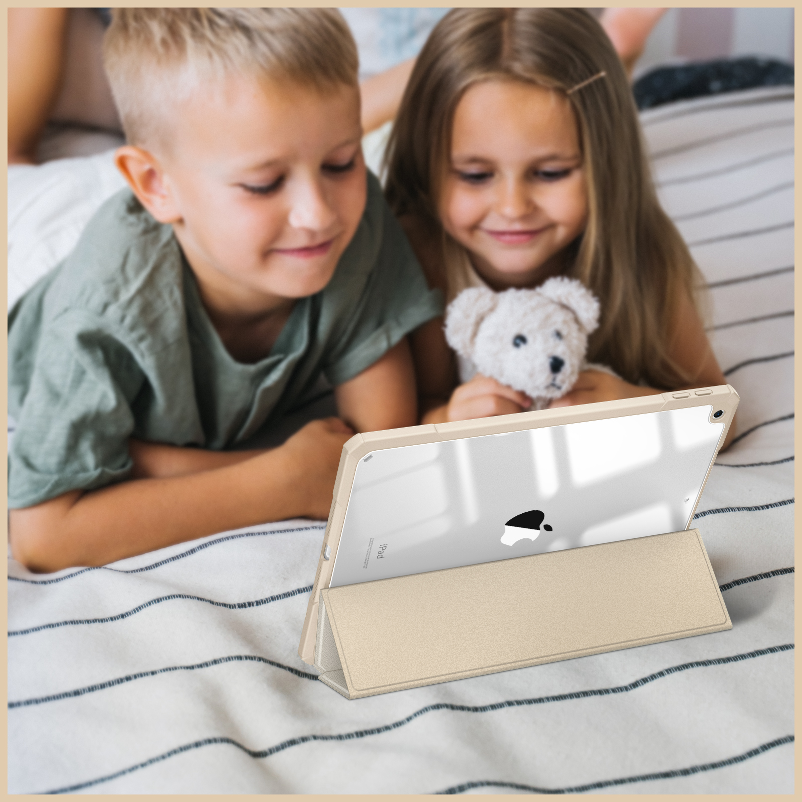 Zwei Kinder schauen auf ein Tablet, das auf einem Bett mit einem Teddybären liegt.