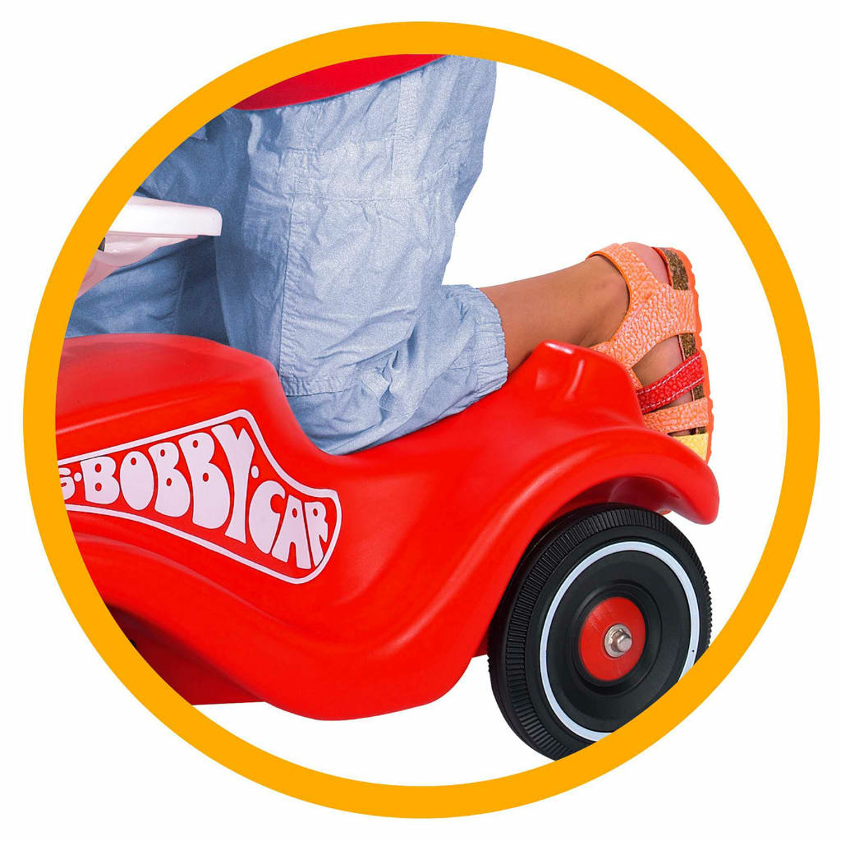 Un bambino è seduto su una Bobby Car rossa. Le gambe del bambino sono visibili. Un cerchio incornicia la scena.