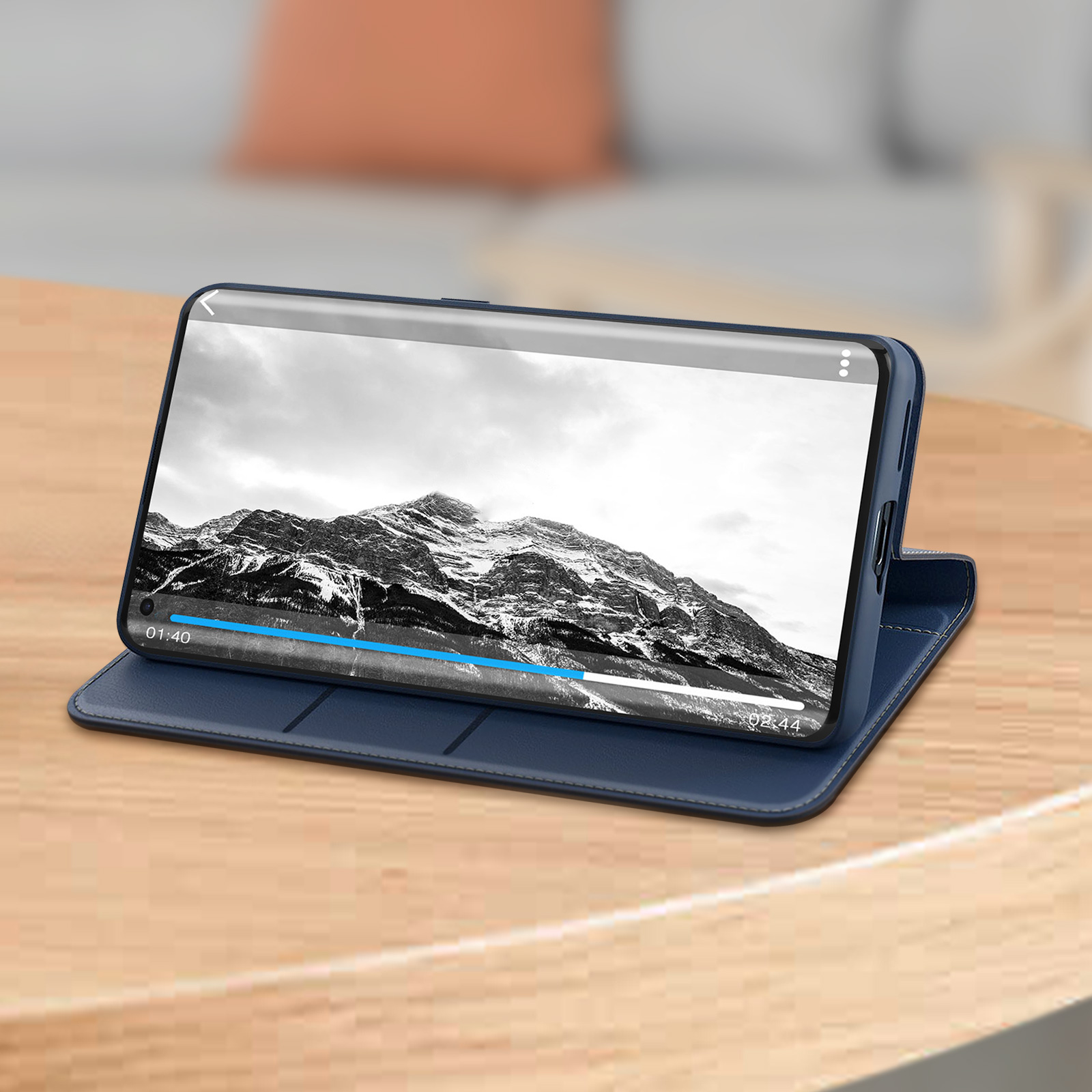 Smartphone zeigt schneebedeckte Berglandschaft, in blauem Gehäuse, auf Holztisch, Sofa im Hintergrund.