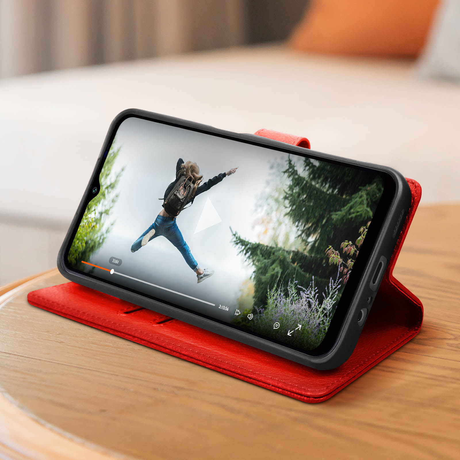 Een telefoon die een video afspeelt, in een rode hoes op een houten tafel. Achtergrond is een bed en een raam.