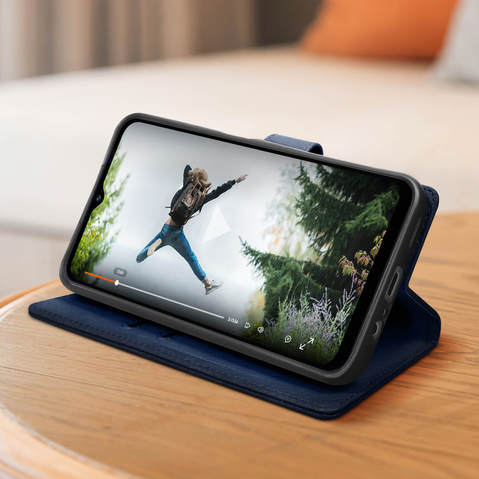 Een telefoon in een blauwe hoes toont een video van een springende persoon. Houten tafel, wazige achtergrond.