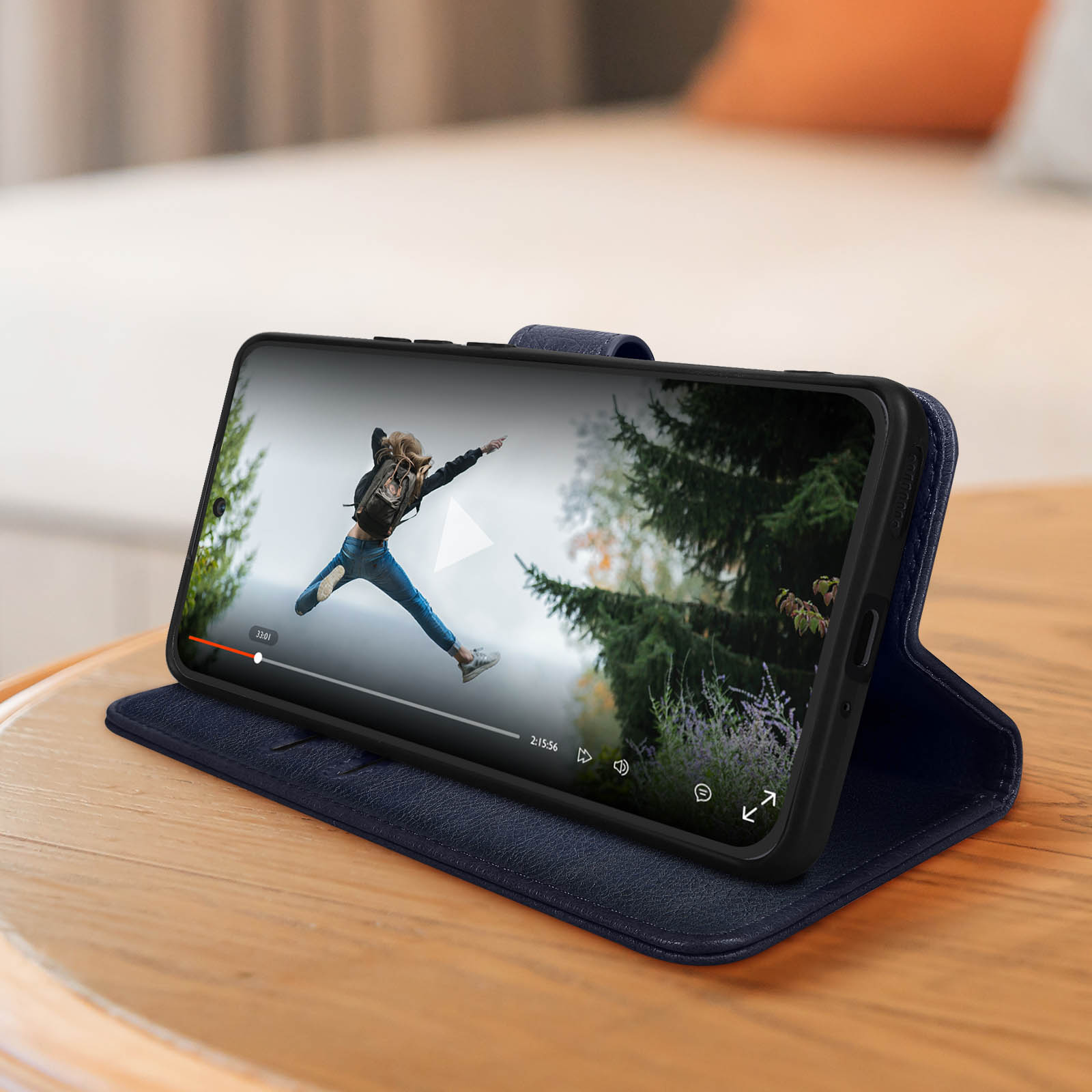 Smartphone avec vidéo d'une personne sautant, dans un étui bleu, sur une surface en bois. Un lit en arrière-plan.