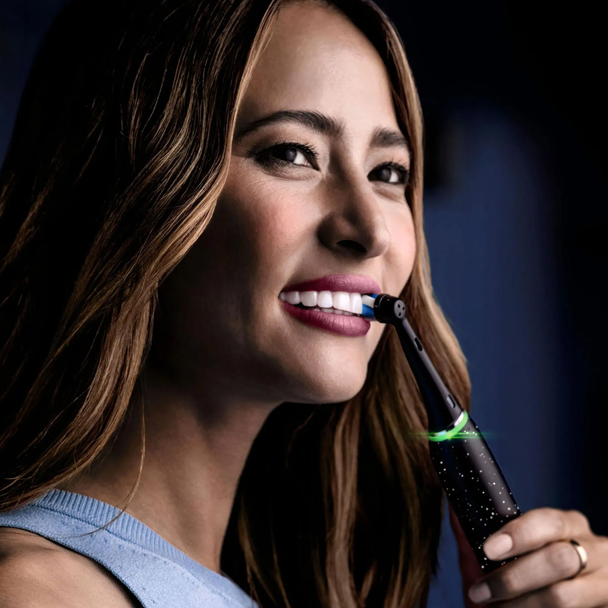 Kobieta uśmiechnięta, trzymająca szczoteczkę do zębów. Czarna szczoteczka do zębów z niebieskim włosiem. Ciemnoniebieskie tło.