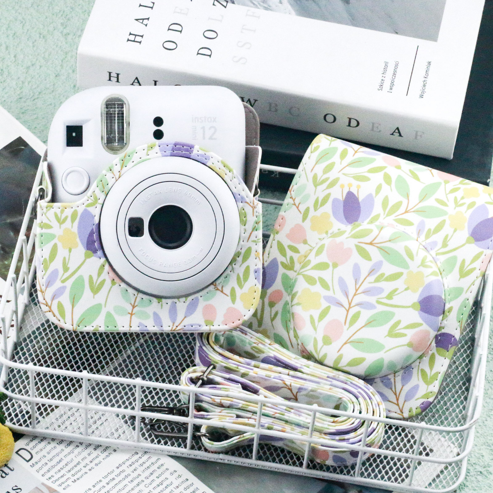 Weiße Instax-Kamera in einer Hülle mit Blumenmuster, mit passendem Riemen und Hülle. Weißer Drahtkorb.
