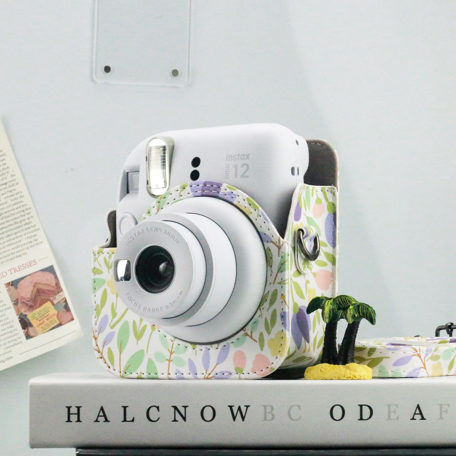 Weiße Instax 12 Kamera in einer floralen Hülle. Sie steht auf einem Buch mit dem Wort "HALCNOW".