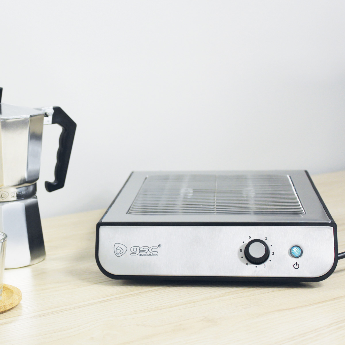 Parrilla eléctrica plateada y negra con dial, cafetera y vaso sobre mesa de madera.