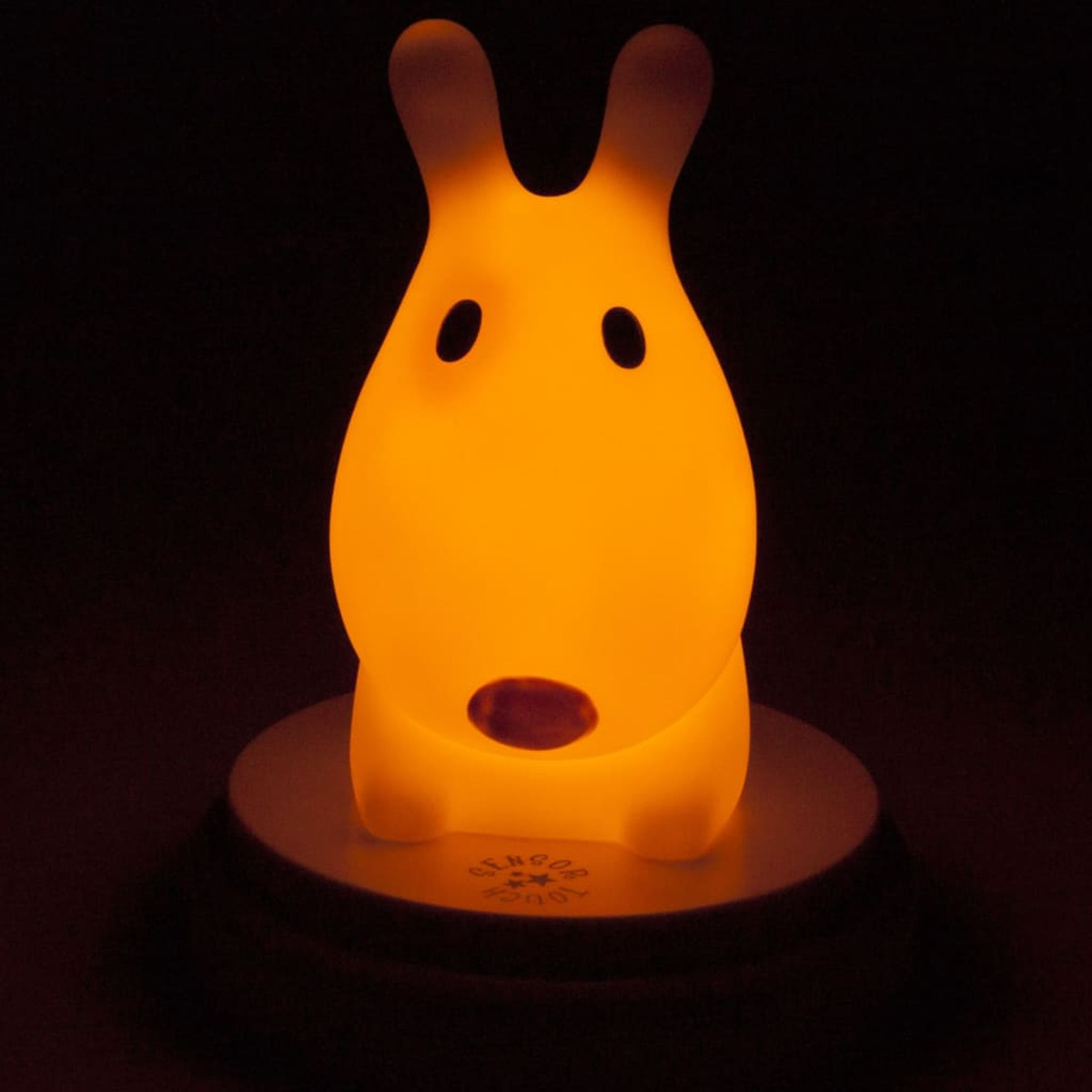 Una luz nocturna en forma de perro brillando en naranja sobre un fondo oscuro, con una base blanca.